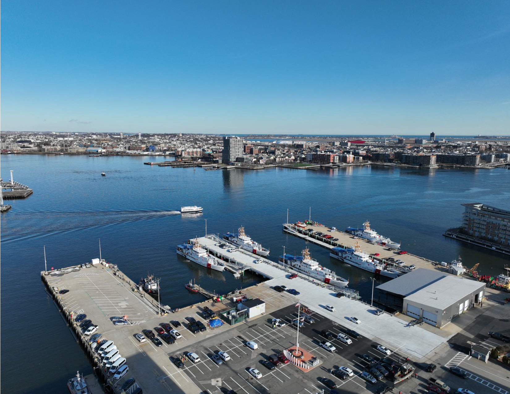 Aerial view of a city harbor with boats and ships docked, and a city skyline in the background under a clear blue sky.