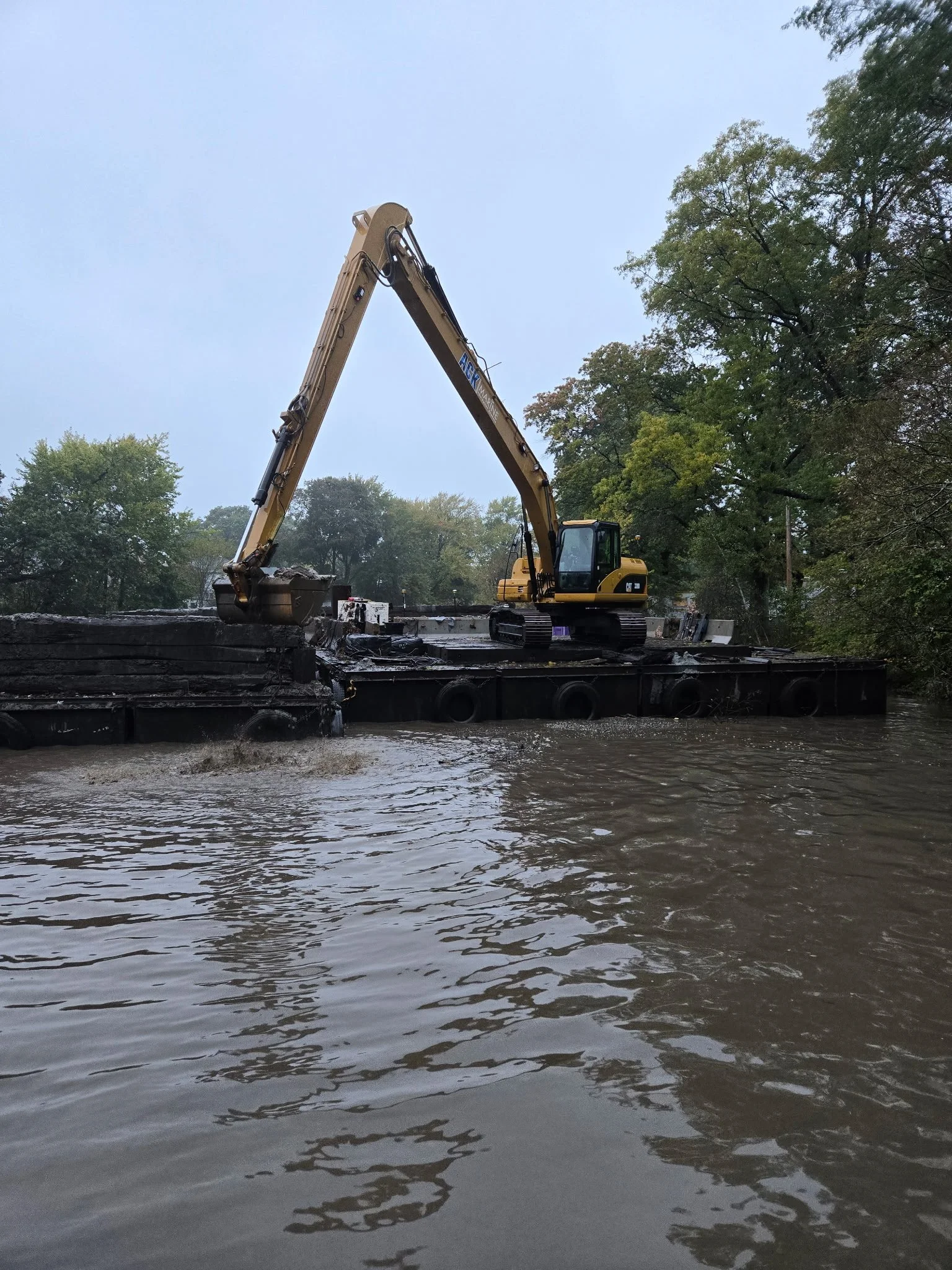 Construction excavator on a barge in a flooded area with trees in the background.
