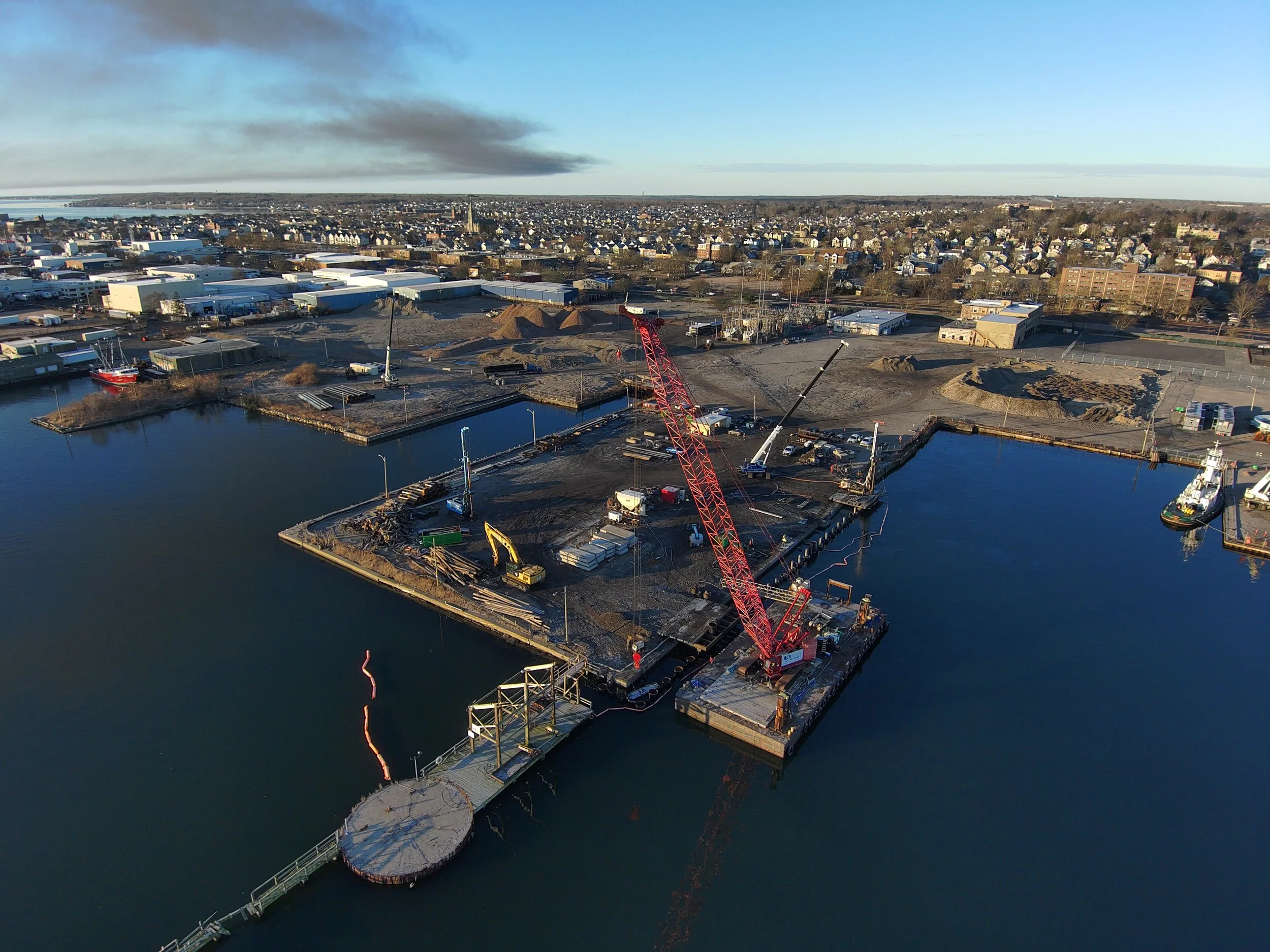 Aerial view of a construction site at a harbor with a large red crane, heavy equipment, and ships docked.