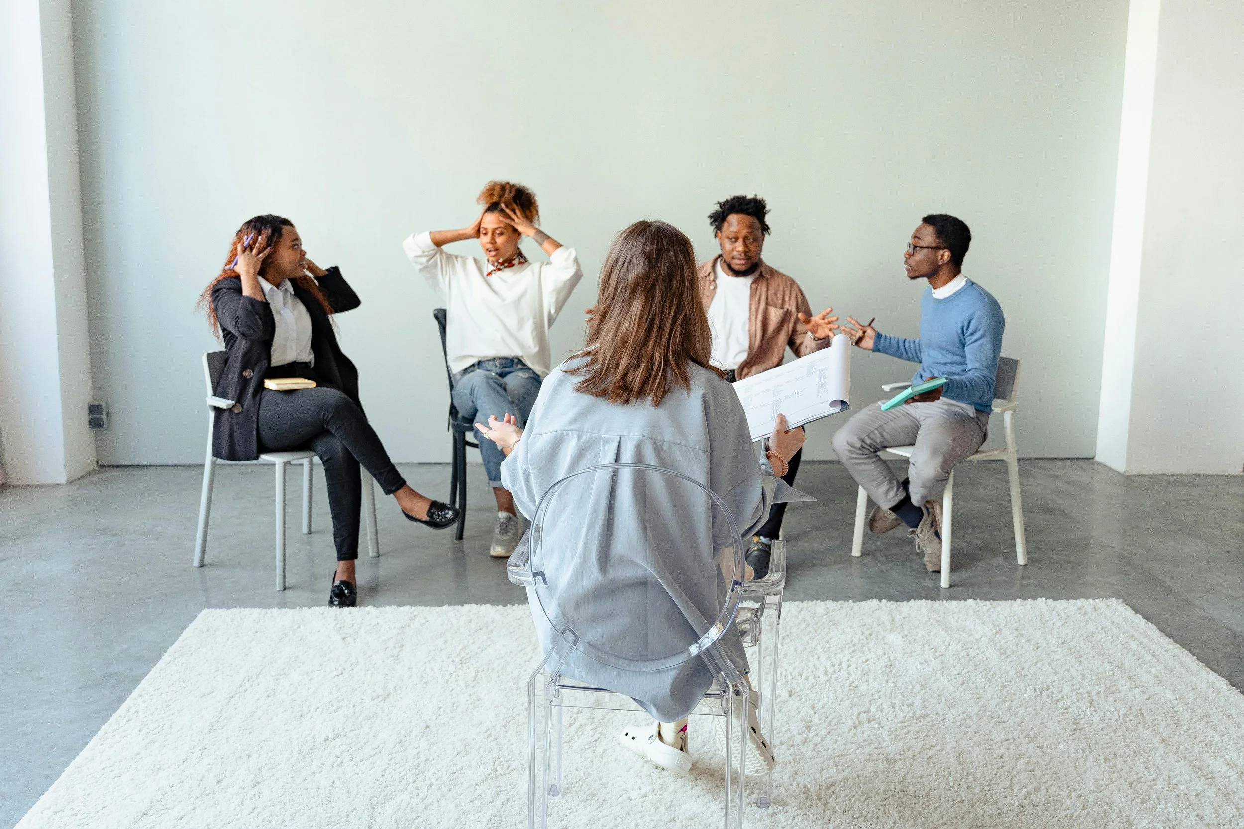 A group of five diverse young adults participating in a discussion or therapy session in a bright, minimalistic room with a white wall and a light-colored carpet.