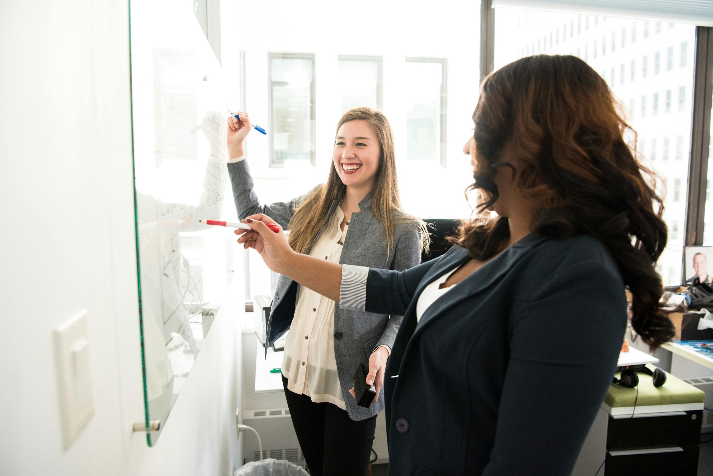 Two women writing on a glass whiteboard in an office, one smiling and holding a marker, the other also writing, with large windows in the background.