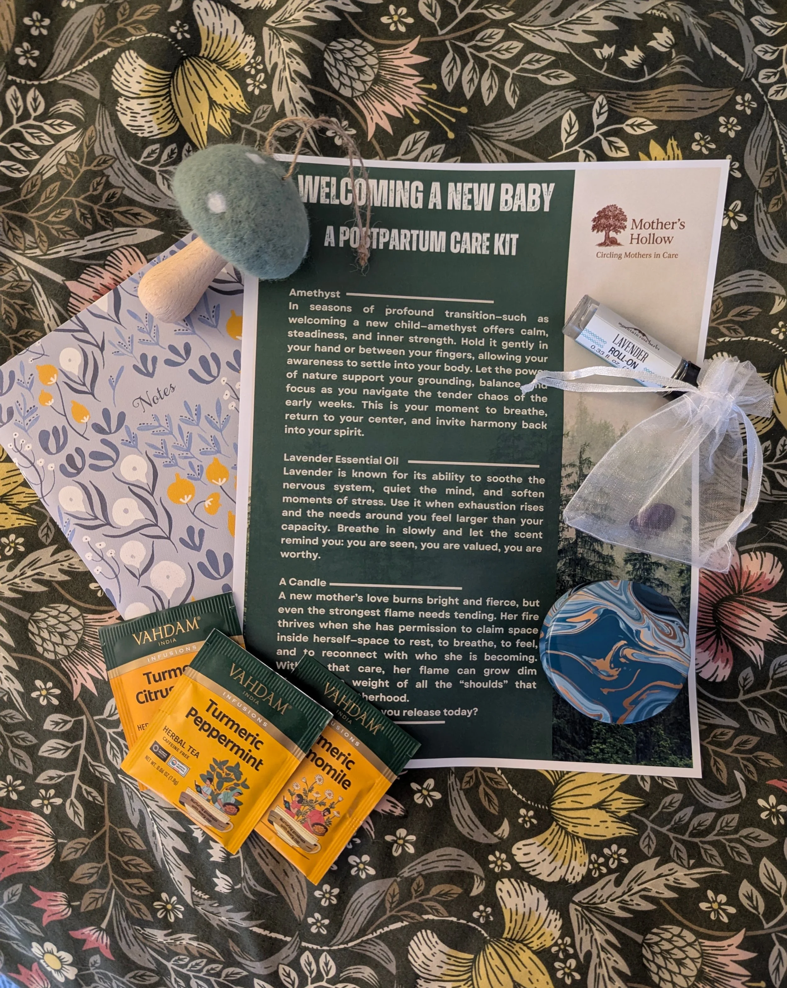 A postpartum care kit for welcoming a new baby, including herbal tea packets, a felted green mushroom, a floral card, essential oil labeled lavender roll-on, a wax candle, and a small organza bag with stones, all arranged on floral patterned surface.