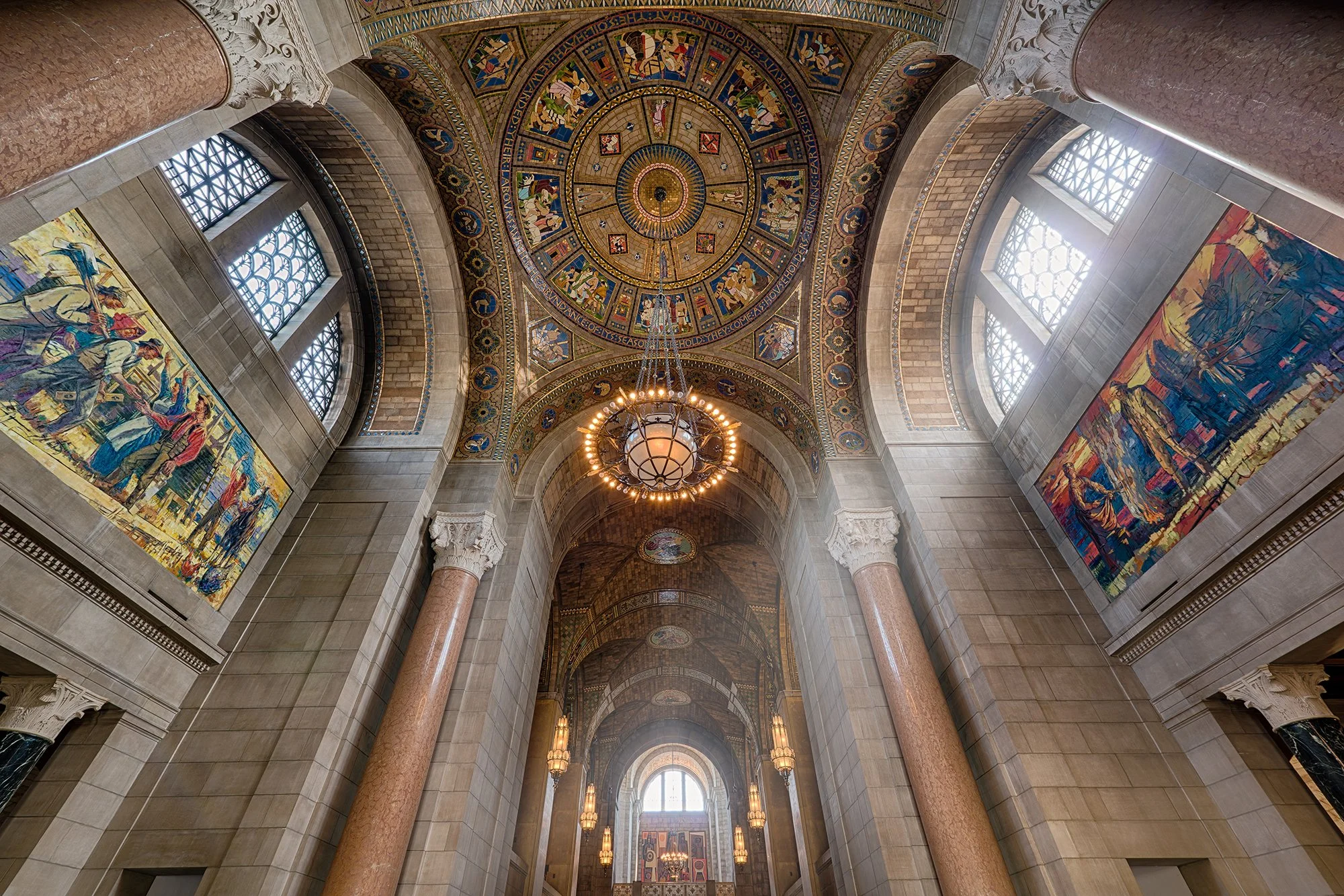 Interior of a grand hall with a high vaulted ceiling decorated with colorful mosaic and a large chandelier hanging in the center. There are large windows with intricate lattice work, tall columns with ornate capitals, and colorful murals on the walls.