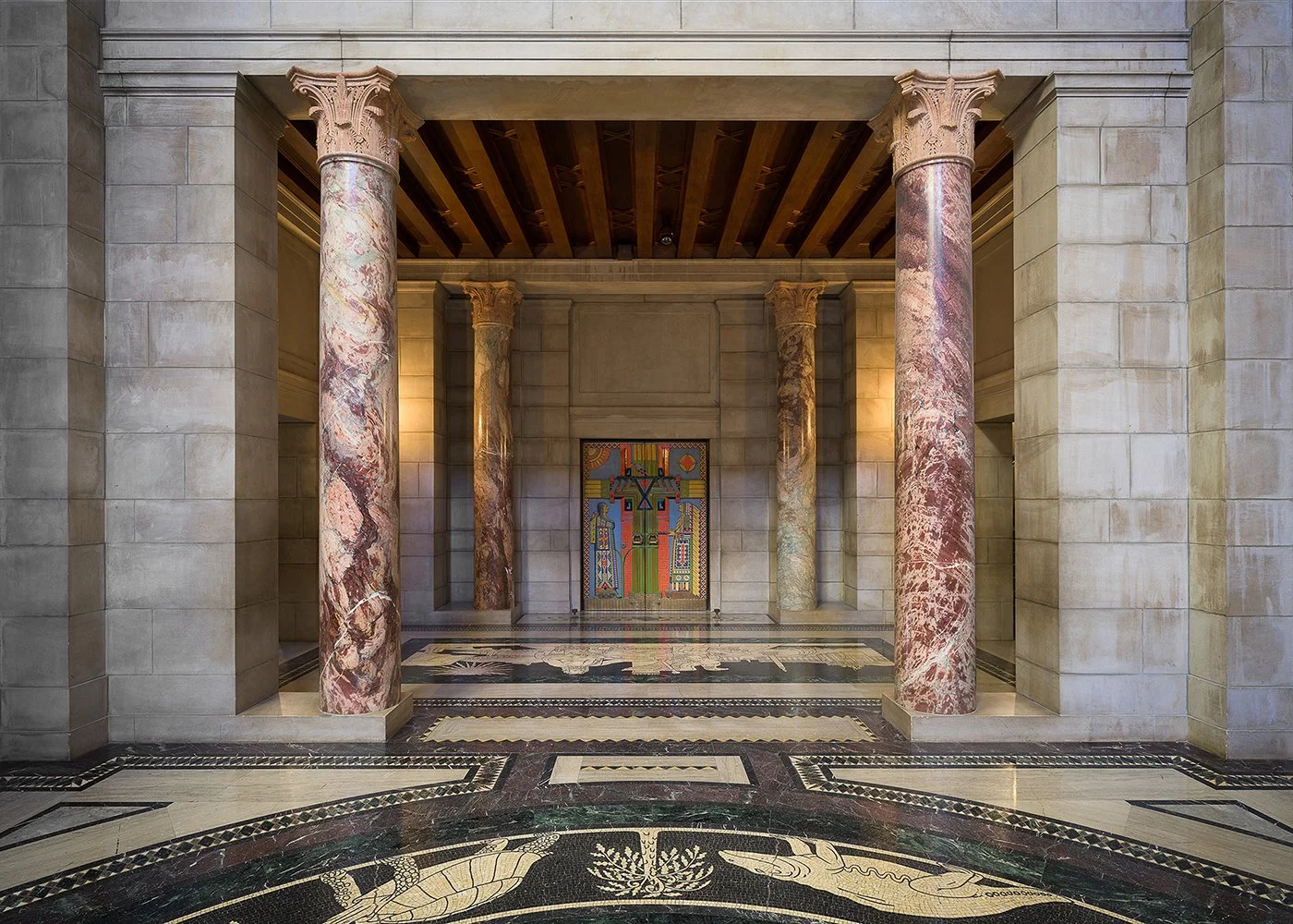 Interior of a building with four classical marble columns, a colorful artwork on a wall, and a patterned marble floor.