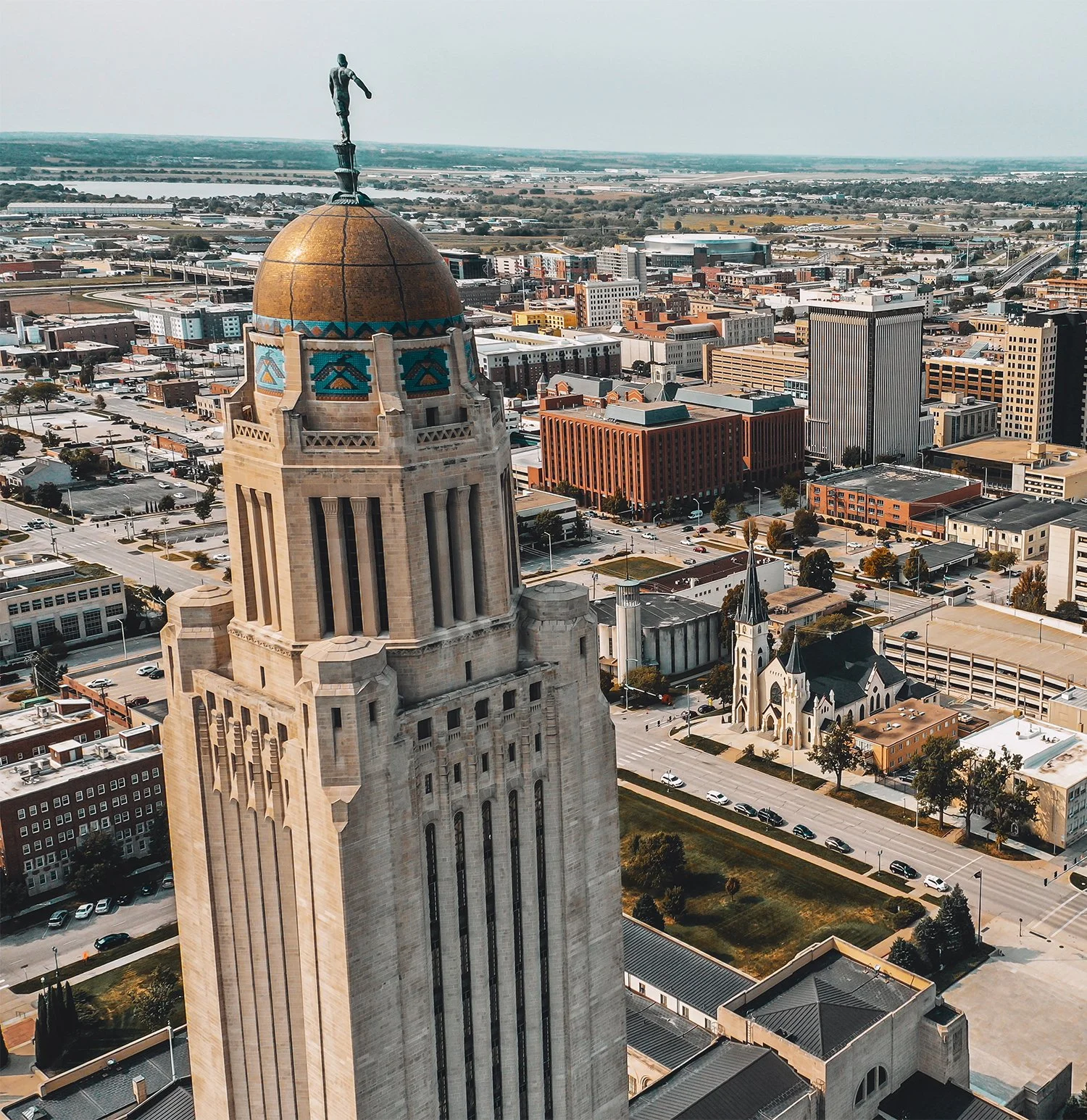 Aerial view of a cityscape with a tall historic tower with a golden dome and a statue on top, surrounded by modern buildings and a church with a steep roof.