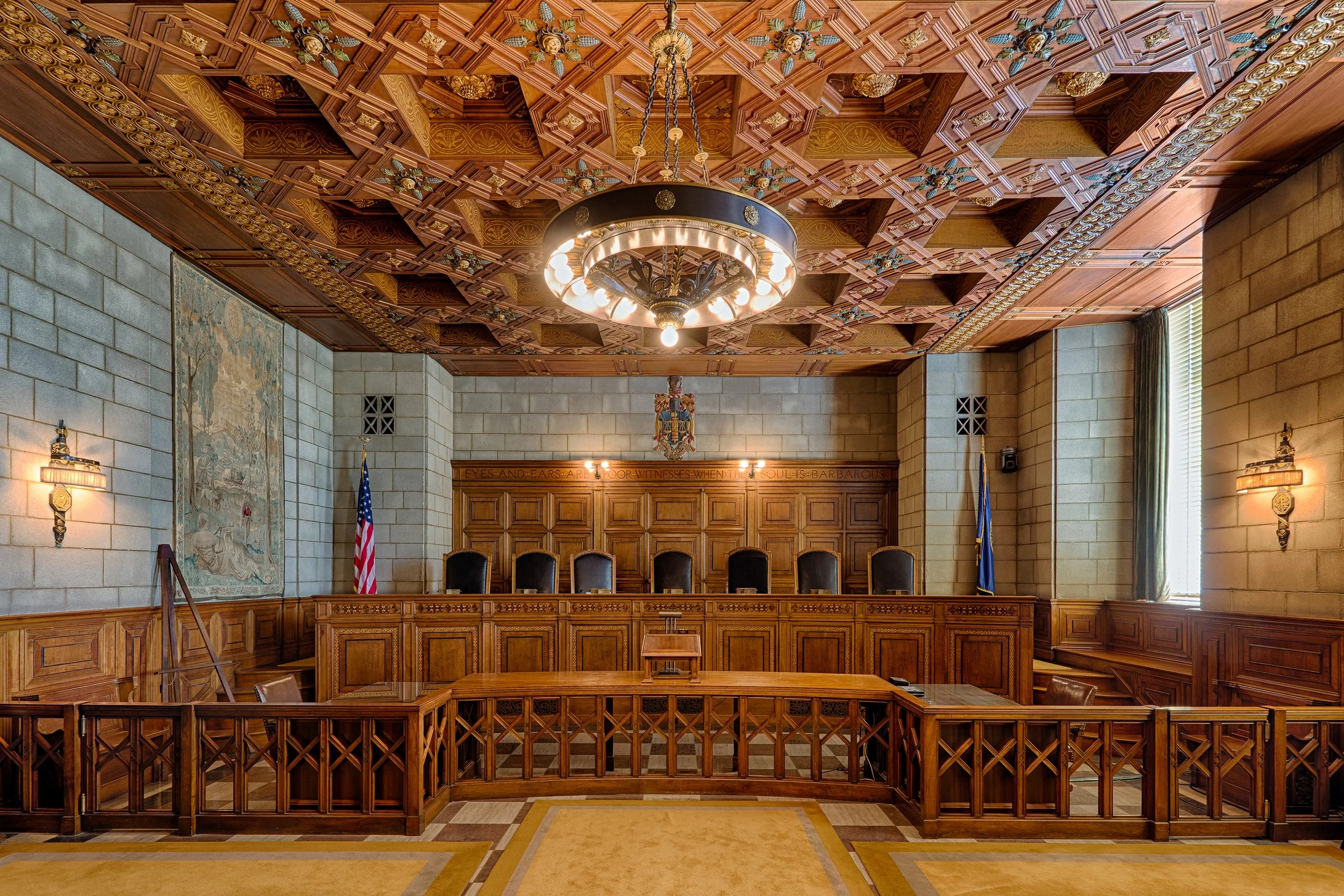 A courtroom with wooden paneling, a high ceiling with decorative woodwork, a chandelier, a row of black chairs behind a wooden bench, American flags, and a large window with curtains.