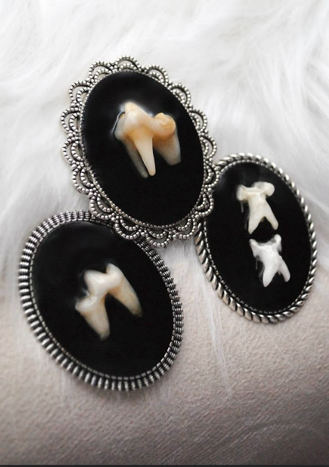 Three vintage-style jewelry pendants with black backgrounds, each holding a small animal tooth or claw, displayed on a white textured fabric.