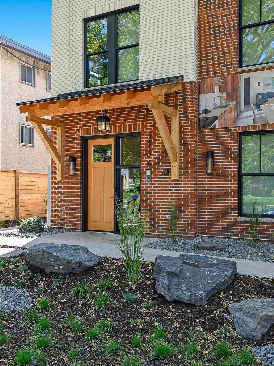Modern brick house entrance with wooden door, large windows, and landscaped front yard with rocks, grass, and plants.