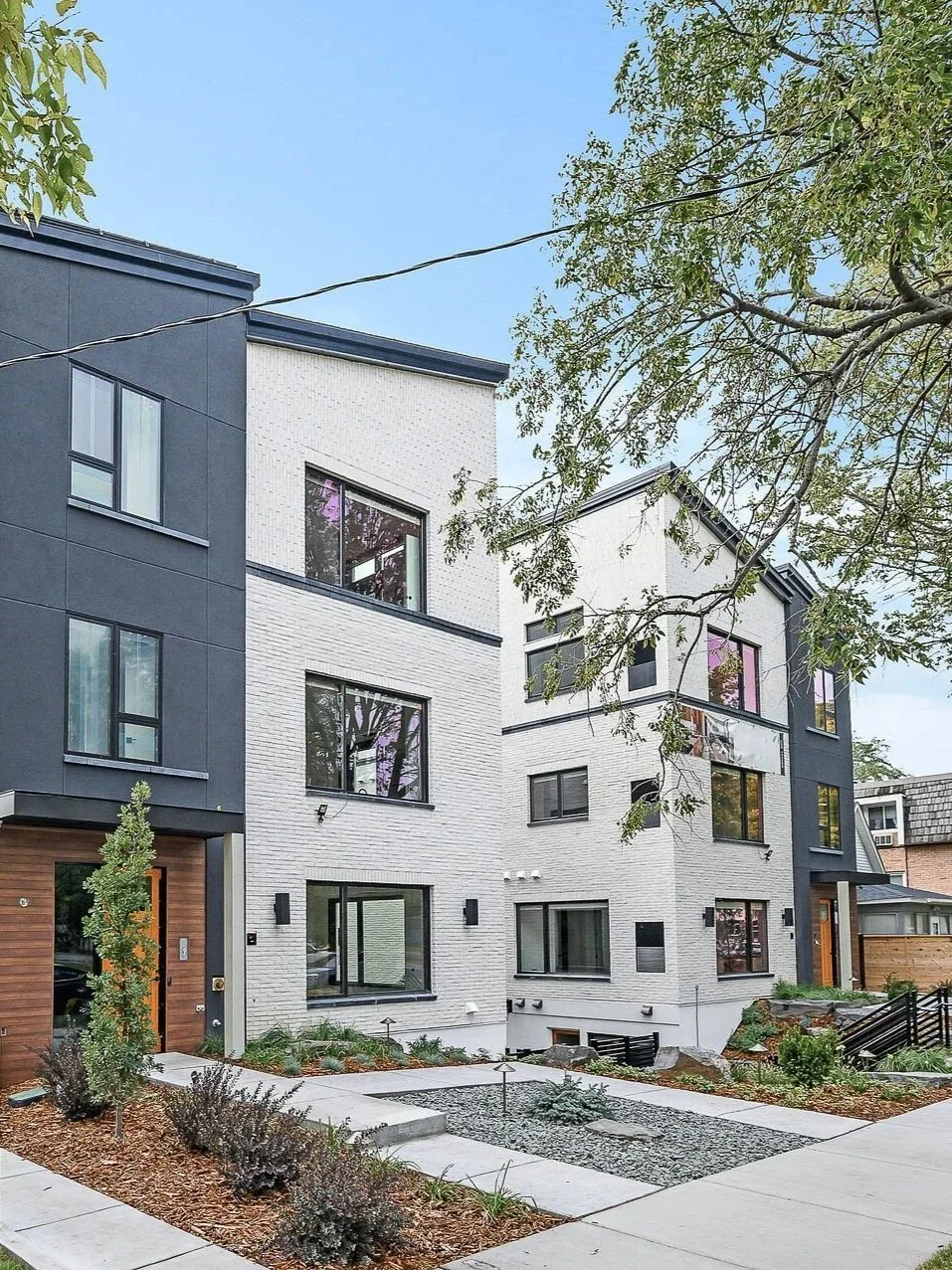 Modern multi-story residential buildings with white brick and dark gray siding, surrounded by landscaped yard with small trees, shrubs, and concrete walkways.