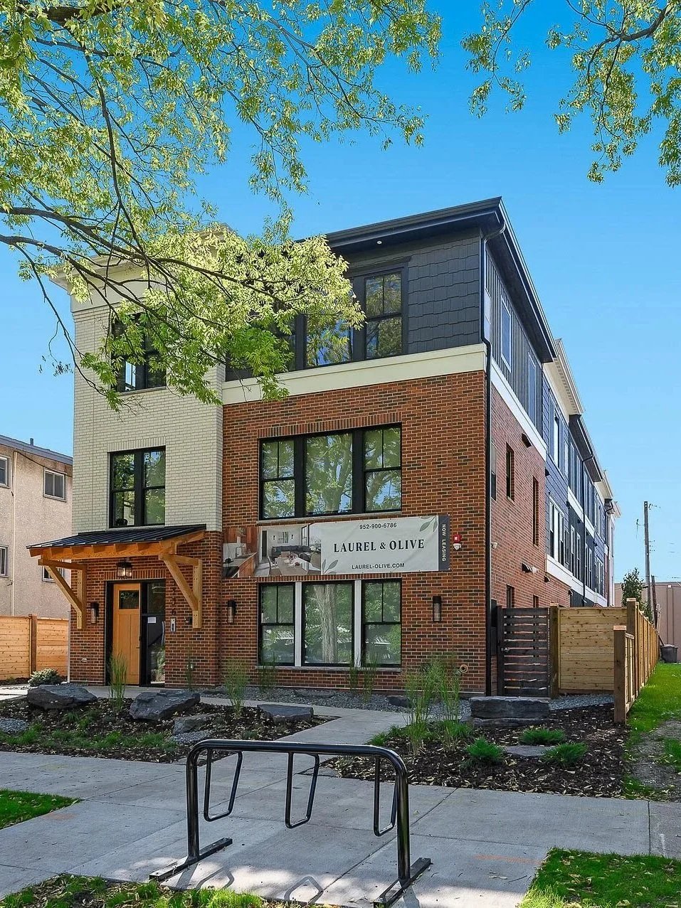Modern four-story brick and dark siding apartment building with large windows, a wooden entrance canopy, and a sign that reads 'Laurel & Olive' in a landscaped area with young plants and a sidewalk.