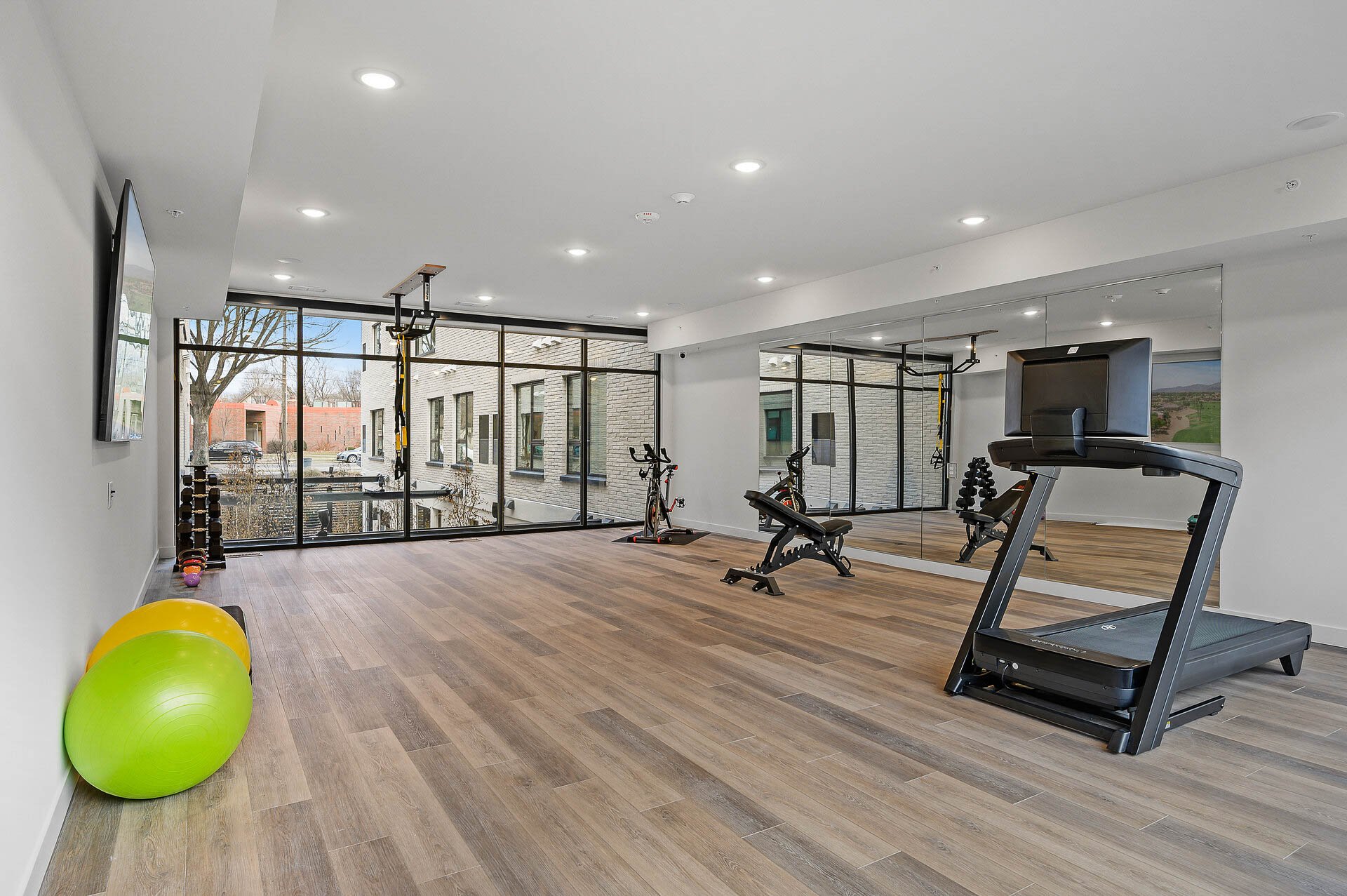 Empty modern gym with wooden floors, large windows, exercise equipment including a treadmill, stationary bikes, dumbbells, and balance balls, with a wall of mirrors.