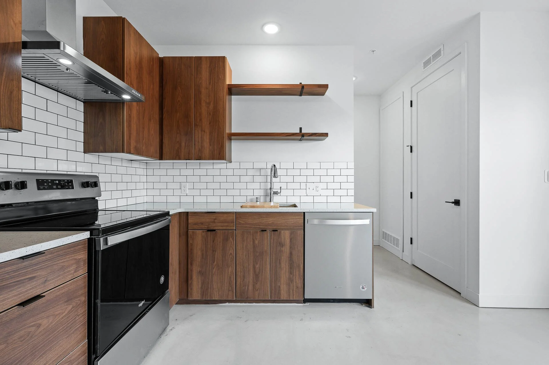 Modern kitchen with white subway tile backsplash, wooden cabinets, a black oven, a stainless steel dishwasher, open wooden shelves, and a white wall with a door.