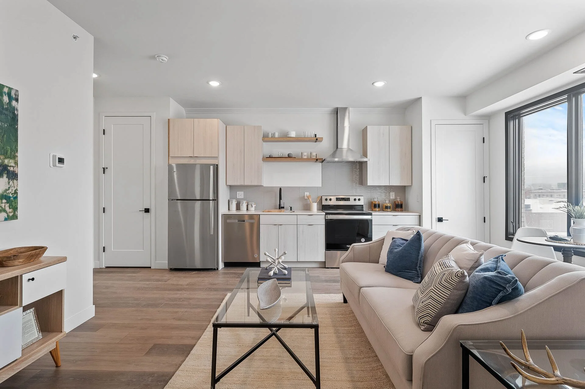 Open-concept living room and kitchen area with a beige sofa, blue and striped throw pillows, glass coffee table, wooden dining table near a window, white cabinetry, stainless steel appliances, and a large window with city view.