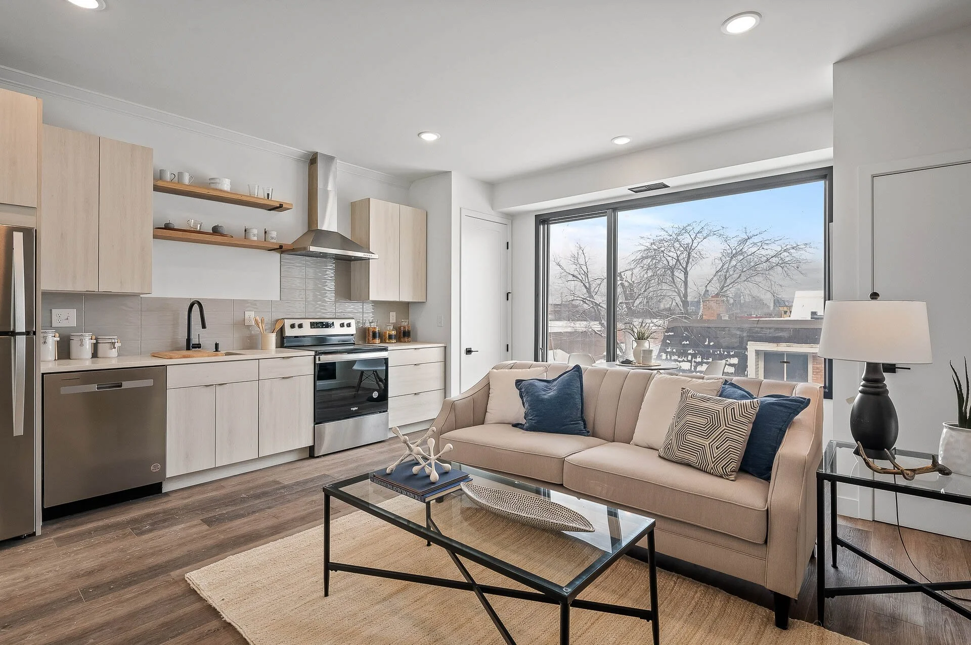 Open-concept living room and kitchen with beige sofa, glass coffee table, large window with view of trees, and light wood cabinetry in the kitchen.
