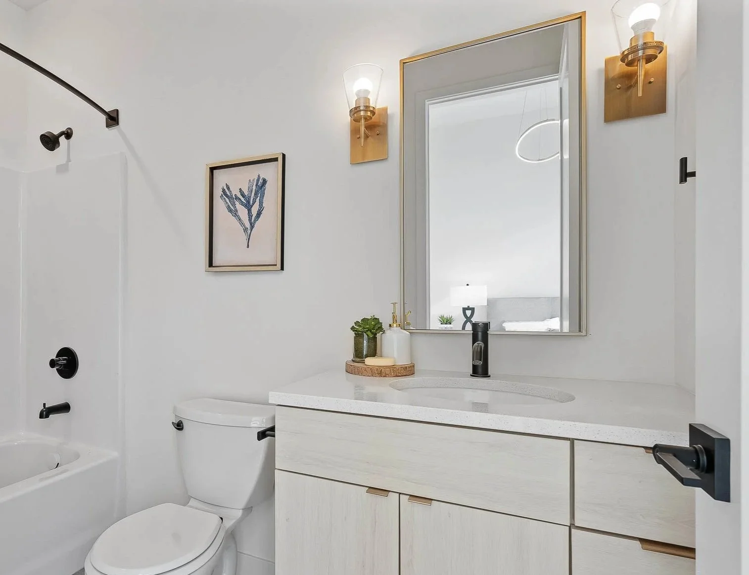 Modern bathroom with white walls, a white toilet, a vanity with a light-colored countertop, a rectangular mirror, and two brass wall-mounted sconces. There is a framed art piece of a plant and decorative items on the countertop.