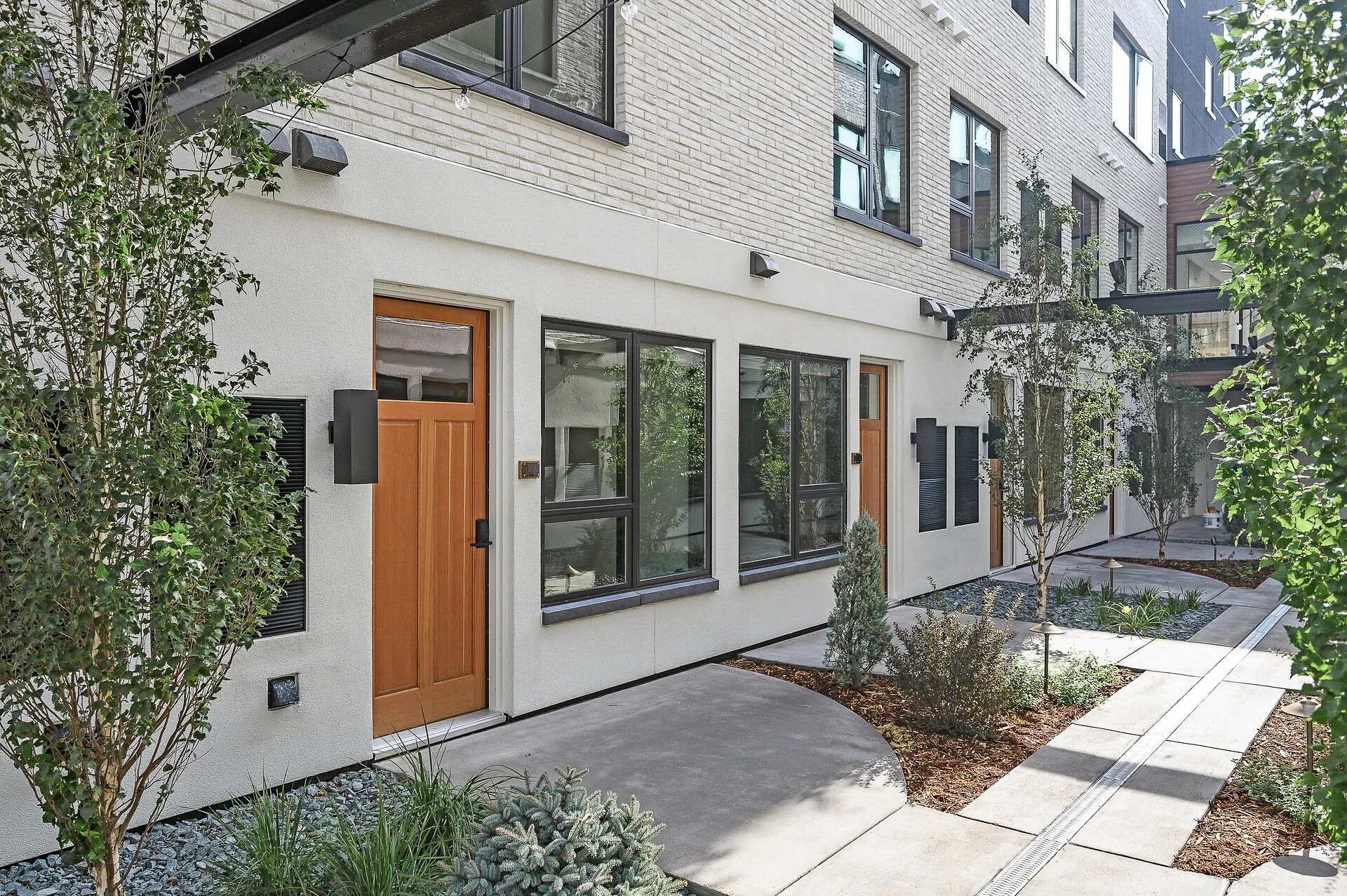 Modern apartment building with multiple doors and large windows, small landscaped garden with trees and plants, concrete walkways, and outdoor lighting.