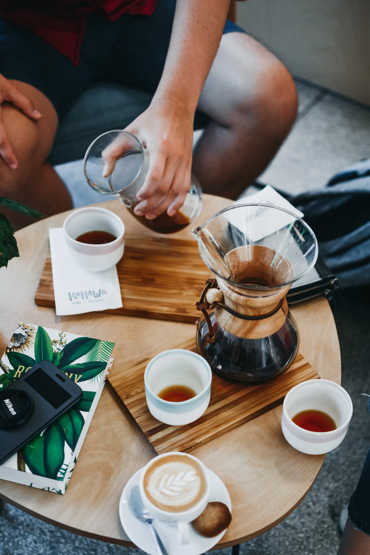 A person preparing pour-over coffee on a wooden table with cups of coffee, a latte, a camera, and a menu.