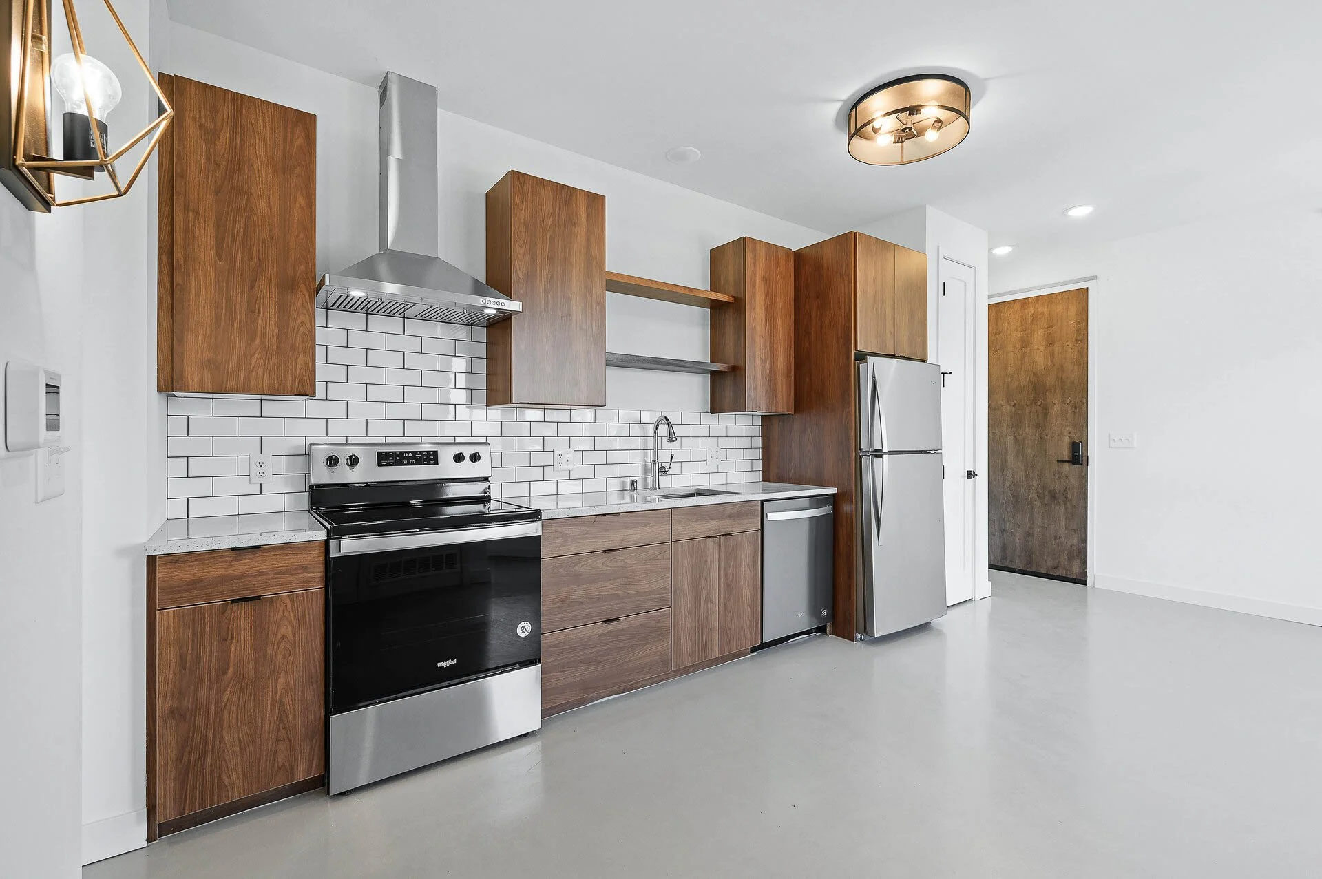 Modern kitchen with white walls, brown wooden cabinets, stainless steel appliances including a stove, refrigerator, and dishwasher, white tile backsplash, and a grey concrete floor.