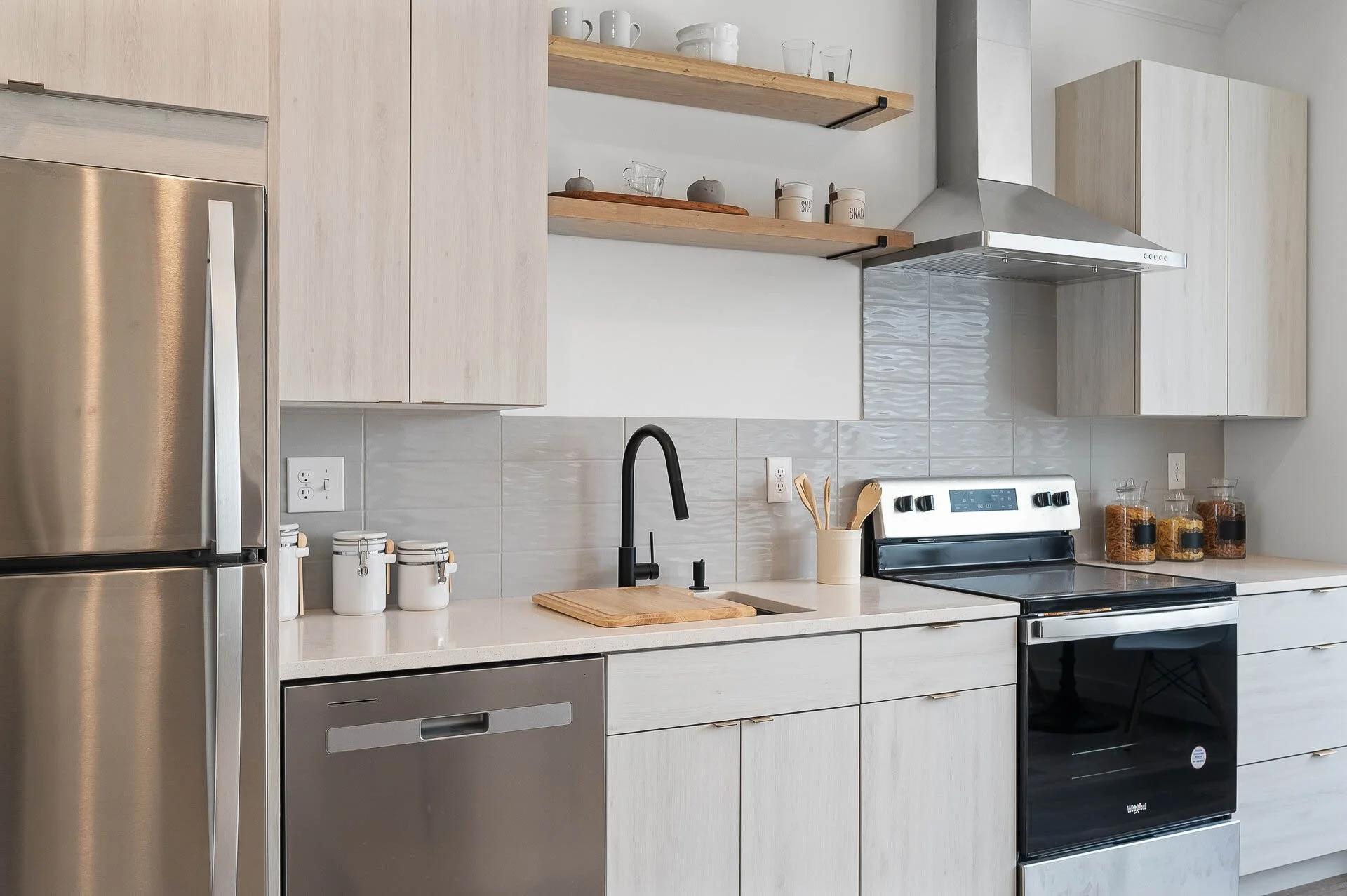 Modern kitchen with beige cabinets, stainless steel refrigerator and dishwasher, black faucet, wooden shelves with glassware and cups, stove with oven, and countertop with jars of dry goods.