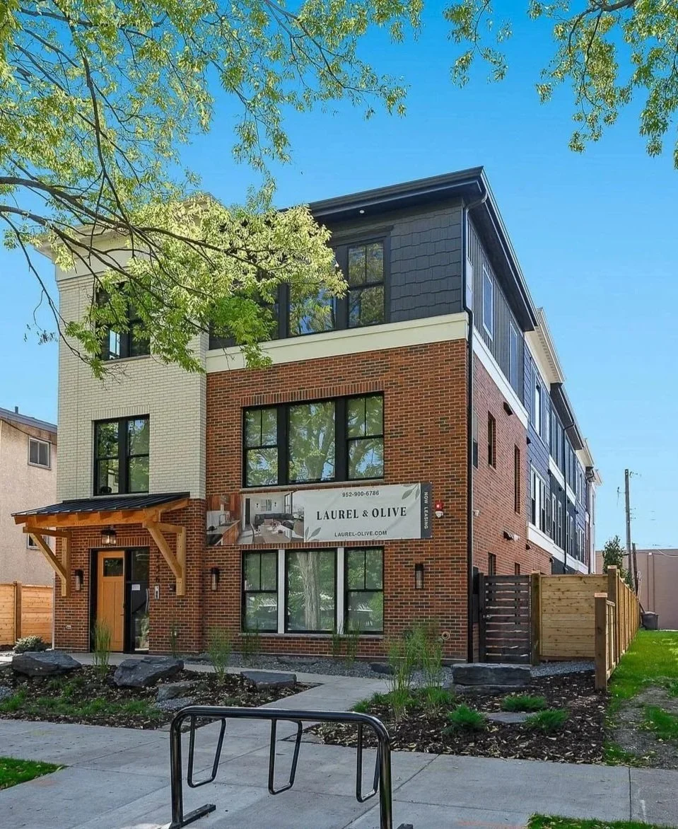 A modern multi-story brick building with large windows and a wooden awning over the entrance. There is a sign on the building that reads "Laurel & Olive" and a sidewalk with landscaped plants and rocks in the front.