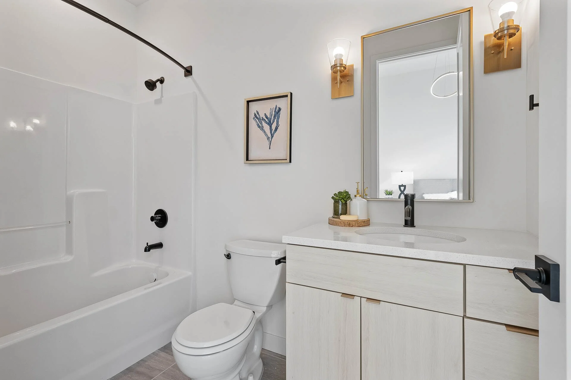 Modern bathroom with a white bathtub, toilet, and vanity with a mirror, wall-mounted lamps, a framed botanical art piece, and decorative items on the sink counter.