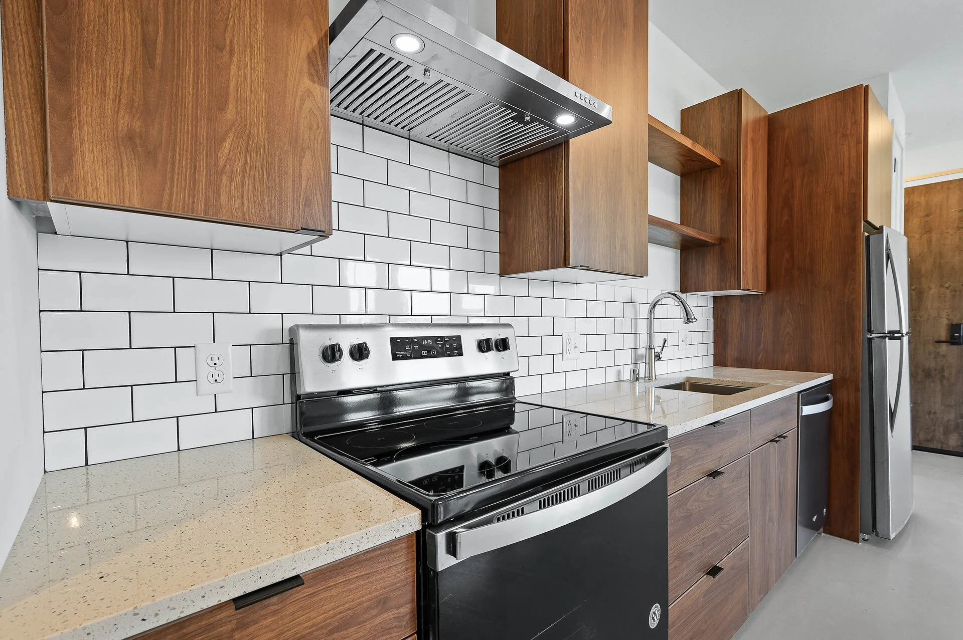 Modern kitchen with white subway tile backsplash, wooden cabinets, black and silver oven, stainless steel range hood, beige countertops, and stainless steel refrigerator.