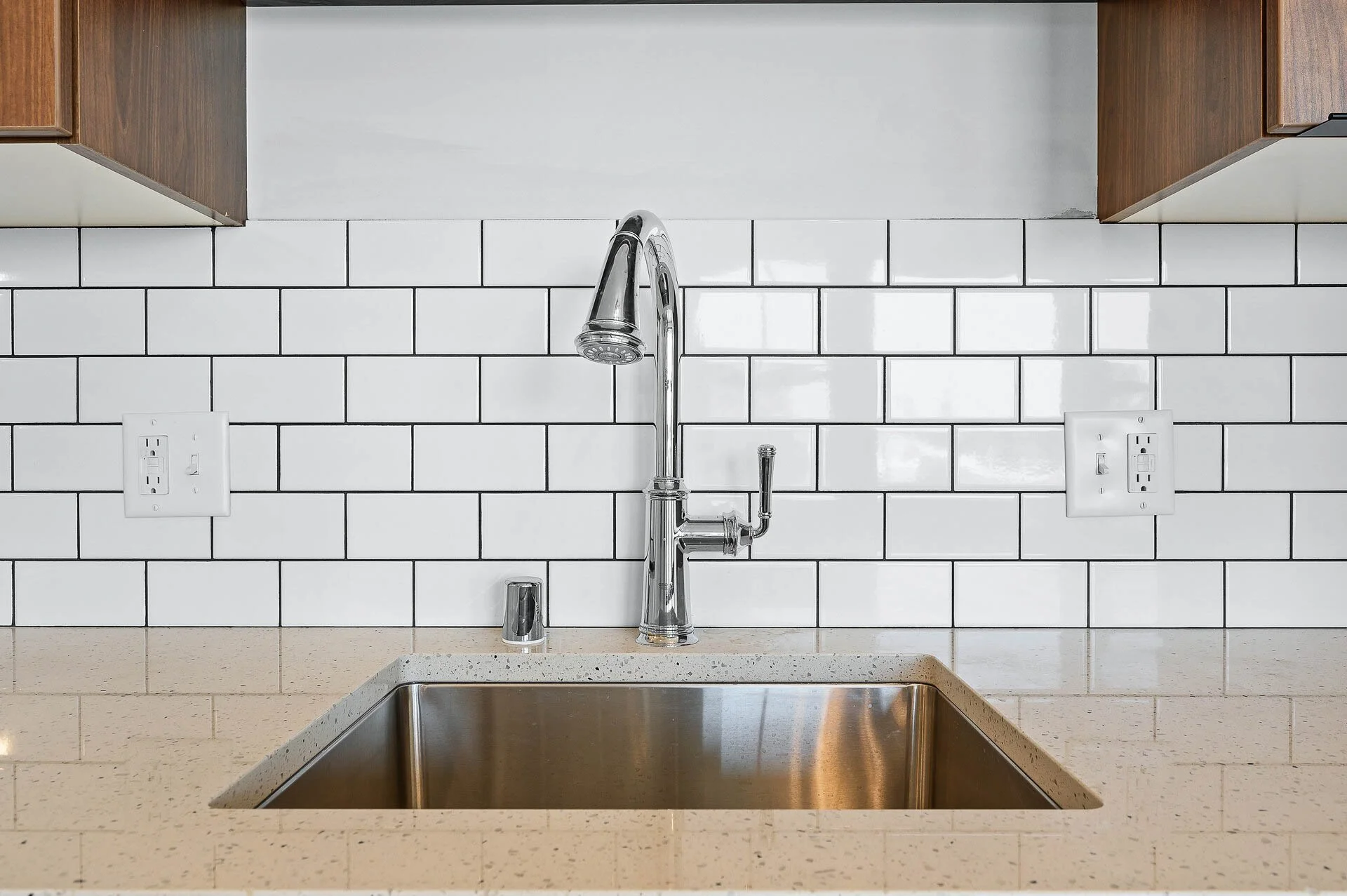 Kitchen sink with a chrome faucet against a white subway tile backsplash and beige countertop. Two electrical outlets are on the tiled wall, with wooden cabinets above.