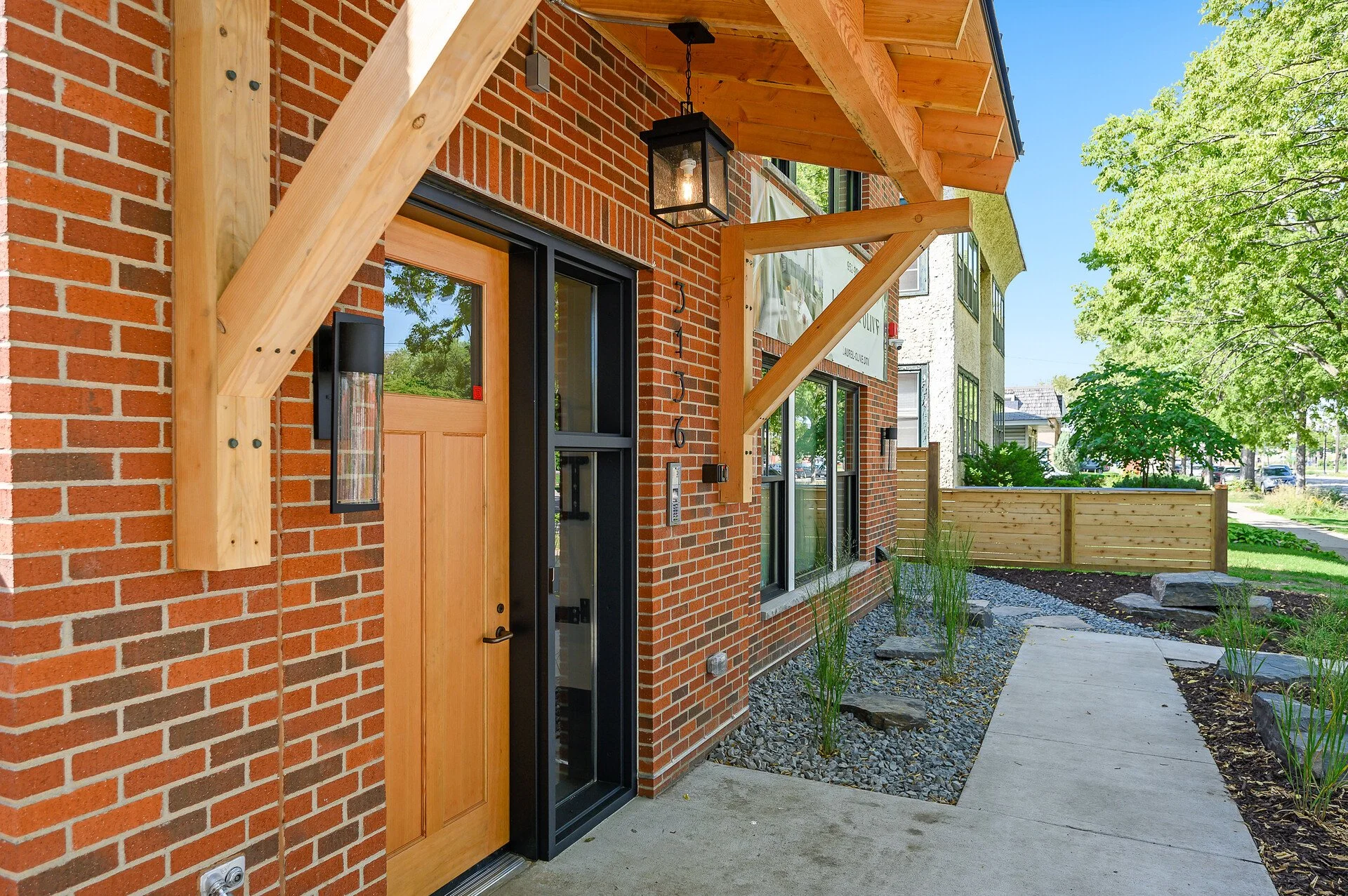 Exterior view of a modern brick building with a wooden door, black metal-framed glass, and a decorative lantern hanging above the entrance. The building has a new wooden awning and trim, and there's a pathway with plants and trees in the surrounding area.