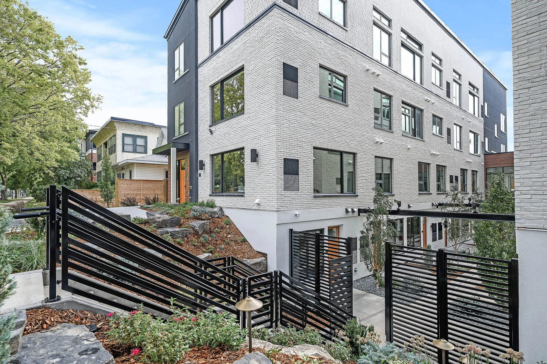 View of a modern multi-story apartment building with white brick and dark accents, surrounded by landscaped greenery, rocks, and a black metal fence on a bright, sunny day.