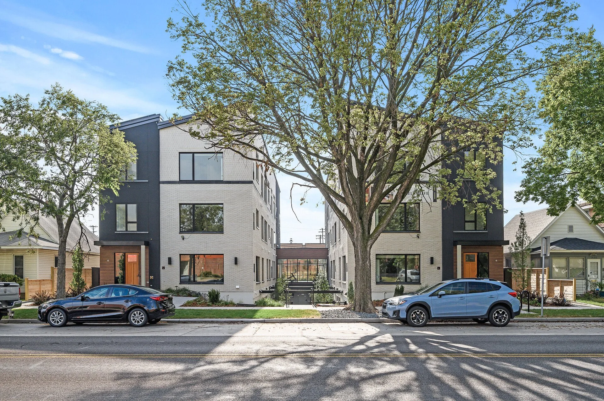 Modern residential building with large windows, two large trees in front, parked cars on the street, and a clear blue sky.
