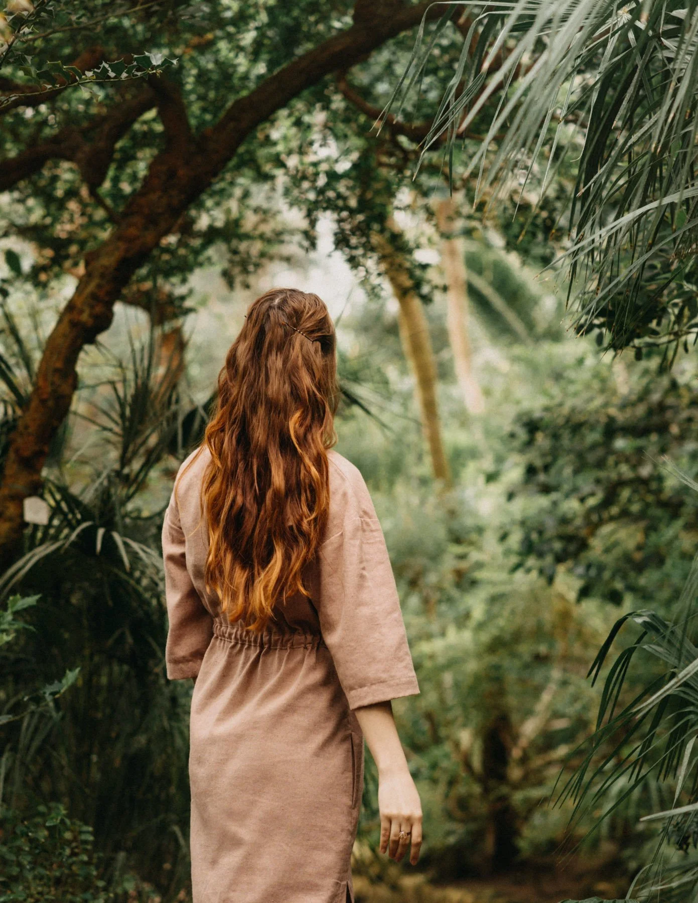 A woman with long, wavy, auburn hair standing in a lush green forest with trees and plants surrounding her.