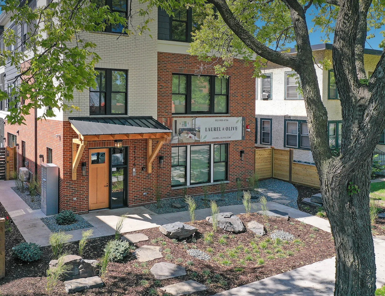 Modern multi-story building with brick facade and large windows, with a landscaped front yard and sidewalk
