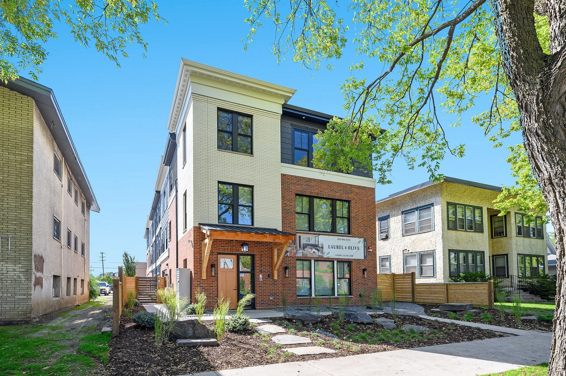 Newly constructed multistory apartment building with brick and siding exterior, black window frames, a small front porch with a wooden awning, landscaped yard, and neighboring residential buildings under a blue sky with green tree foliage.