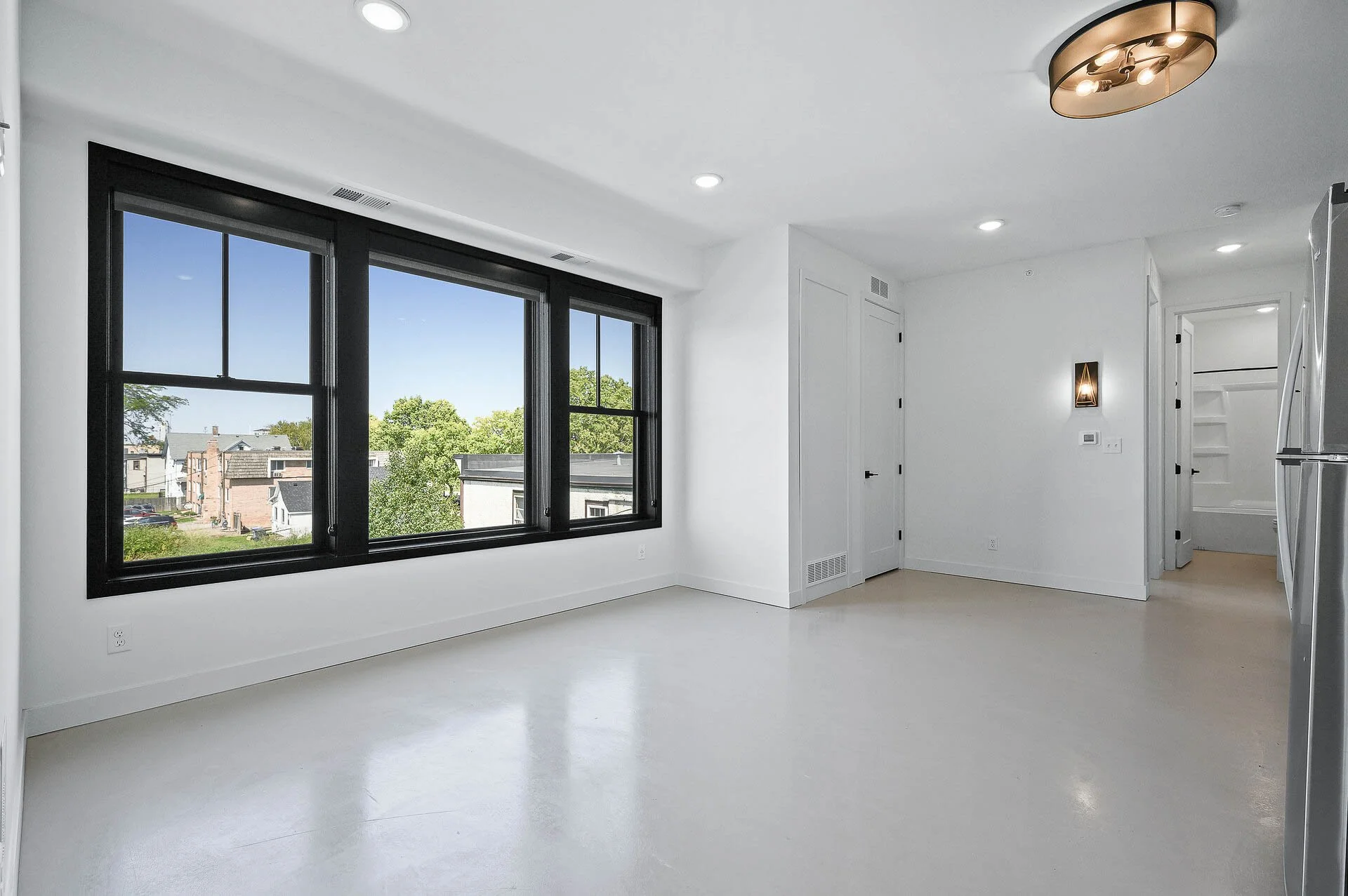 An empty room with large black-framed windows, white walls, beige flooring, ceiling lights, and a view of a residential neighborhood outside.
