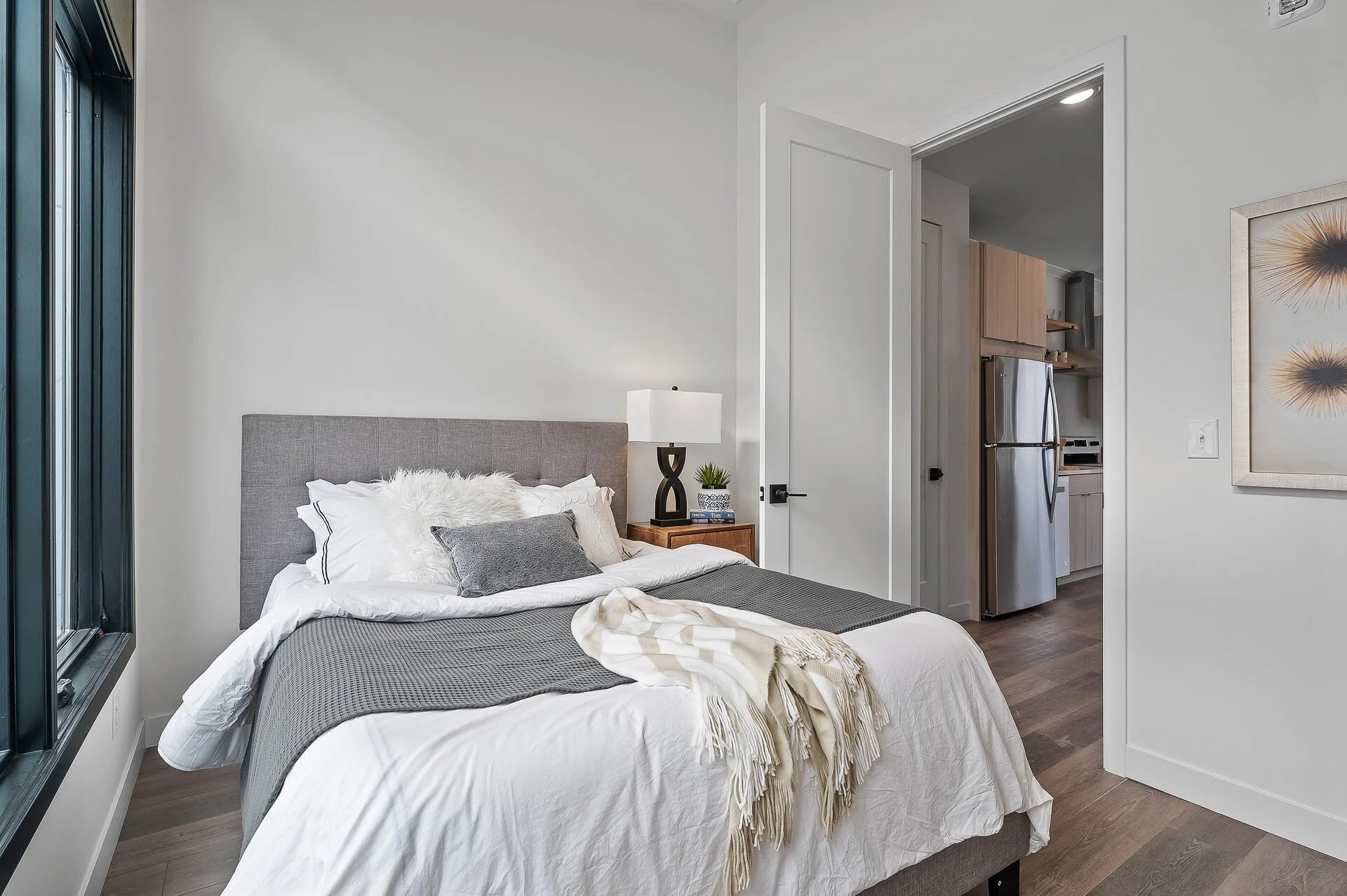 A bedroom with a bed featuring gray, white, and fluffy pillows, placed next to a window with dark frames. A nightstand with a decorative lamp, small plant, and books is beside the bed. An open door reveals a kitchen with a stainless steel refrigerato