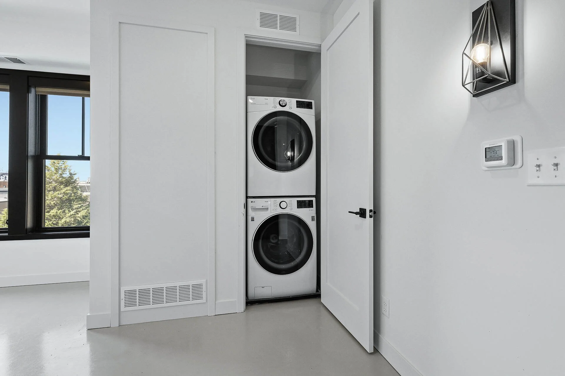 A laundry closet with a stacked white washing machine and dryer, partially open door, white walls, a wall-mounted light fixture, a thermostat, a power outlet, and a window with a view outside.