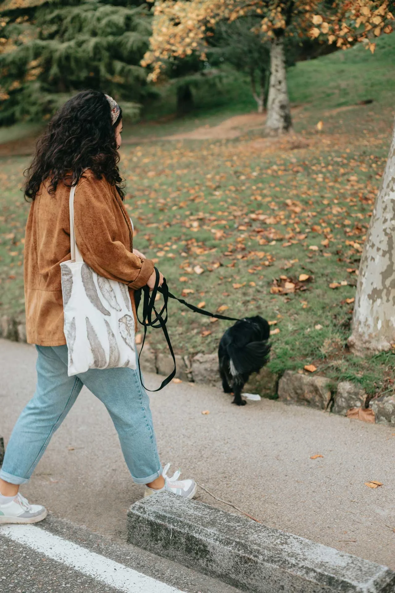 A woman with curly hair wearing a brown jacket, light blue jeans, and sneakers walking a black dog in a park during autumn. The woman carries a tote bag with a dog pattern. Fallen leaves are scattered on the ground, and trees with orange and green leaves are in the background.
