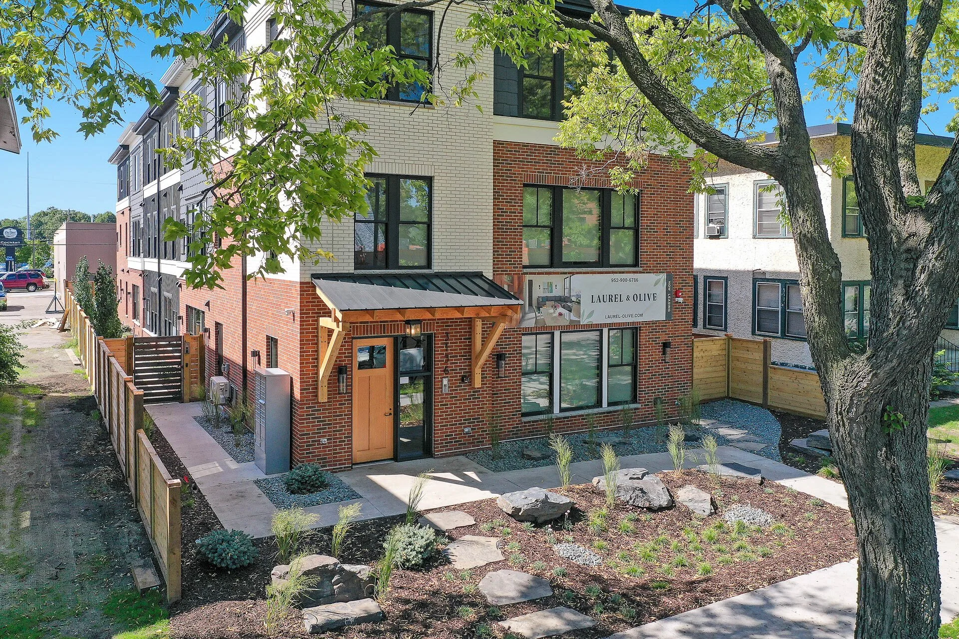 New three-story brick apartment building with a sign that reads 'Laurel & Olive' and a URL 'laurel-olive.com', surrounded by a landscaped yard with stepping stones, plants, and rocks, and shaded by a large tree in the foreground.