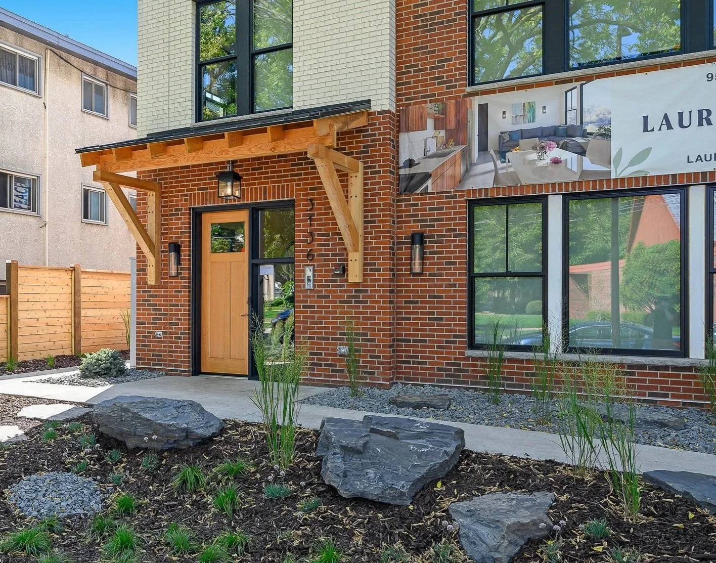 Modern brick apartment building with a wooden front door, large windows, and a small landscaped front yard with rocks and plants.