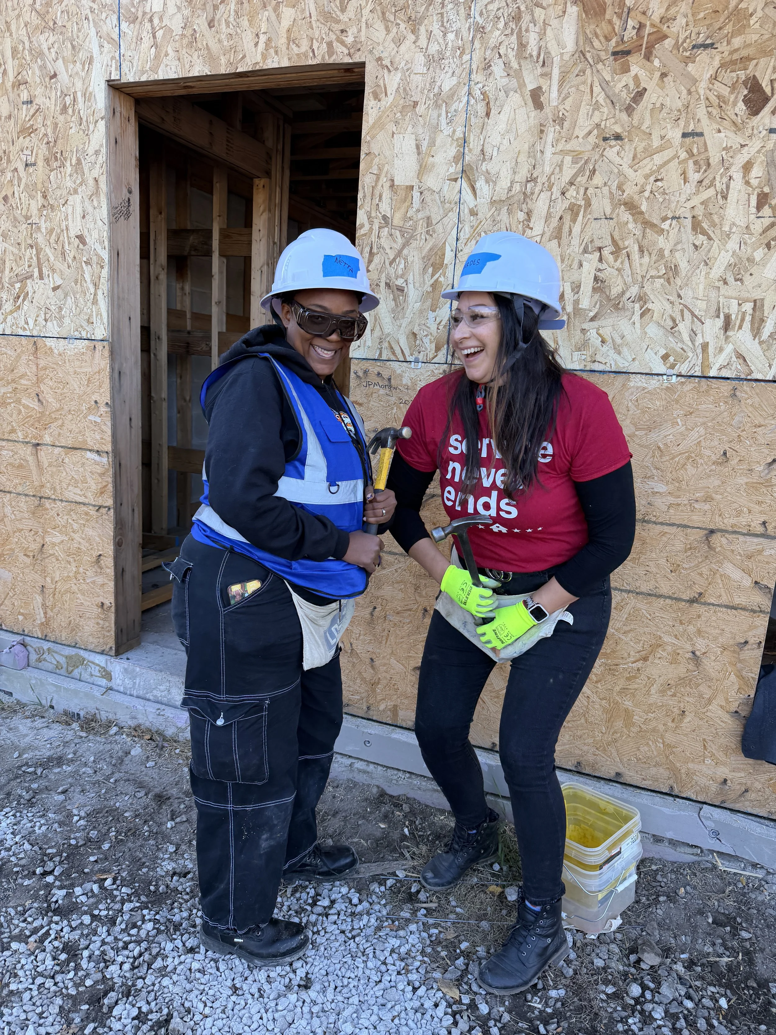 Two women veterans in construction gear, smiling and holding hammers, standing in front of a house under construction for Habitat for Humanity.