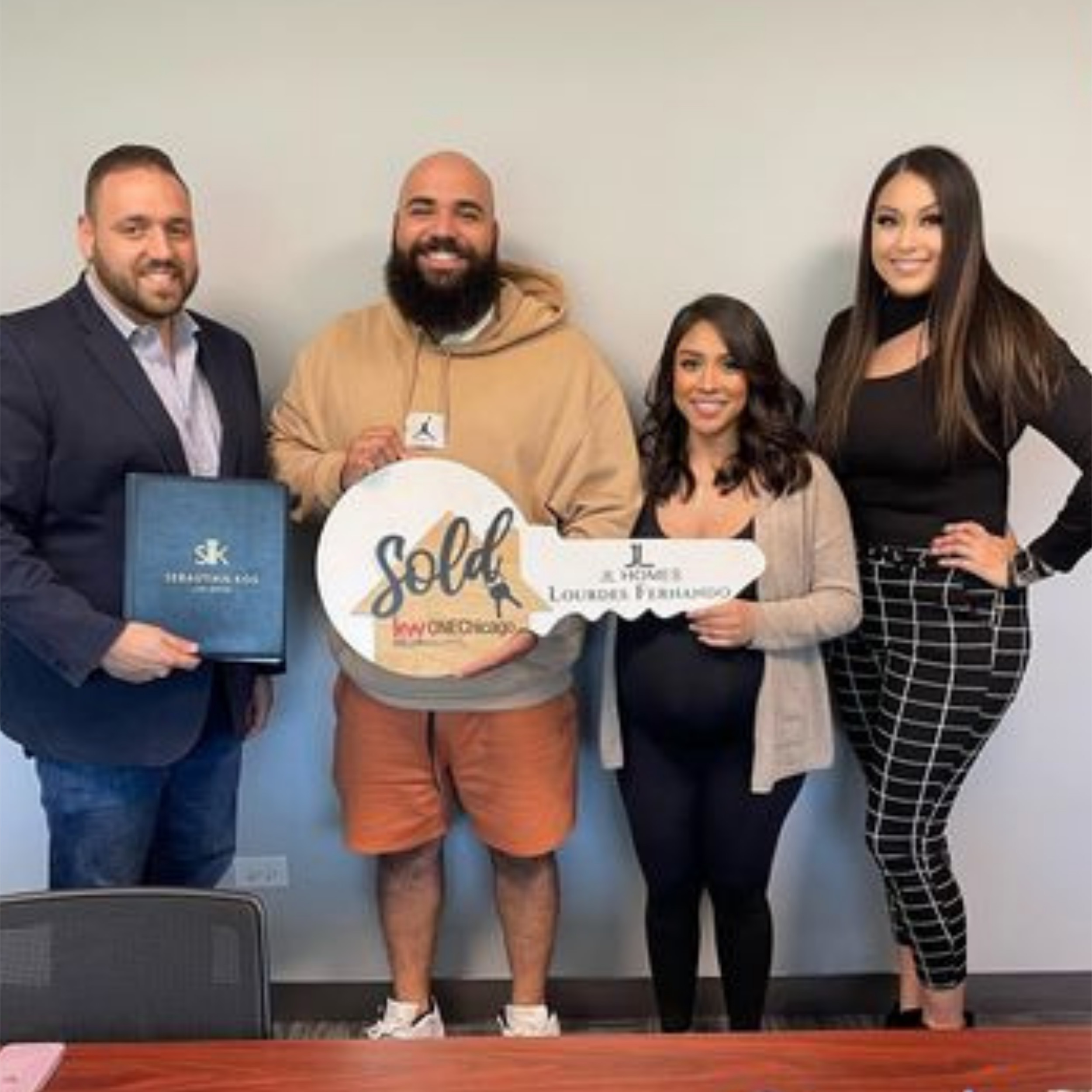 Group of four people standing indoors, holding a key shaped sign that says "Sold" and blue closing folder, celebrating a successful home purchase.