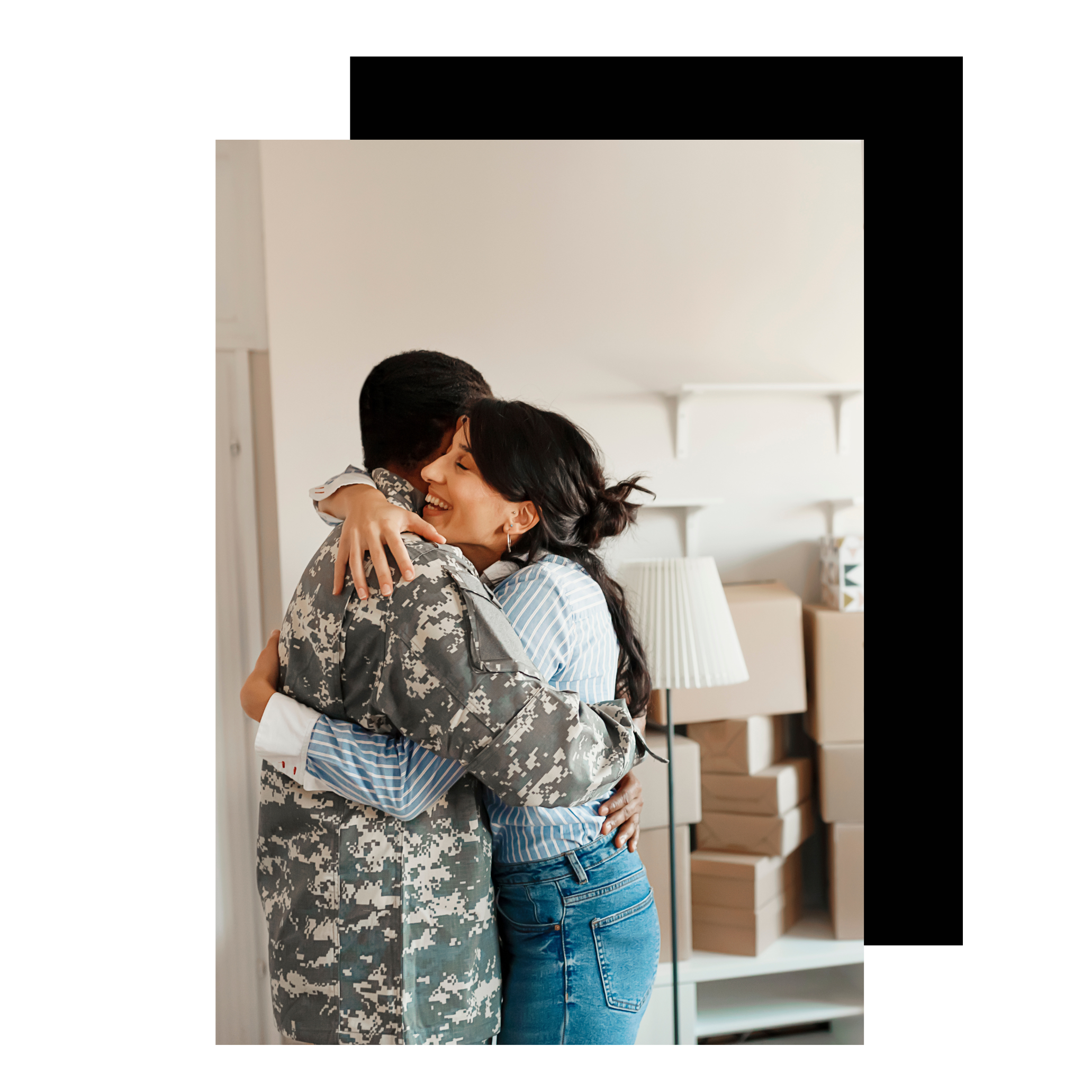 Two women hugging, one in military uniform and the other casually dressed, in a room with boxes and a lamp in the background.