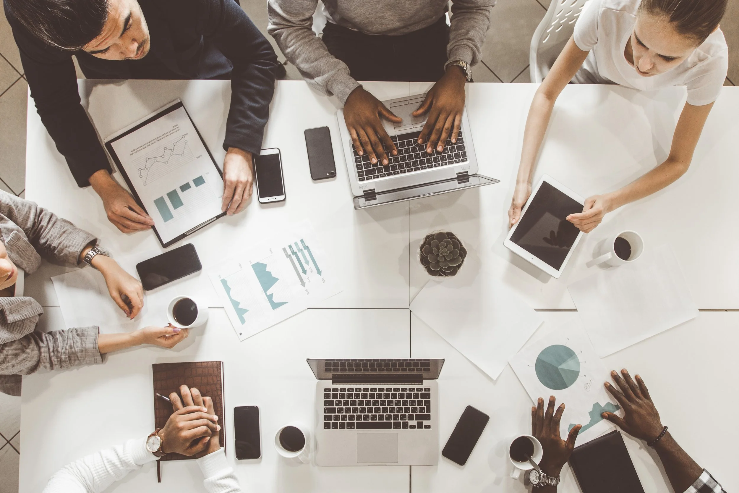 Top-down view of a diverse group of six people at a meeting table, using laptops, tablets, smartphones, and analyzing printed charts, with cups of coffee on the table.