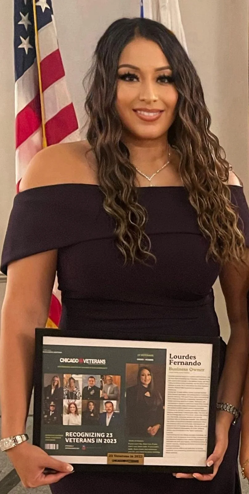 A woman, Lourdes Fernando, with long curly brown hair wearing an off-the-shoulder black dress, standing in front of an American flag, holding a framed article from Chicago Veterans recognizing her as a 2023 veteran award winner.