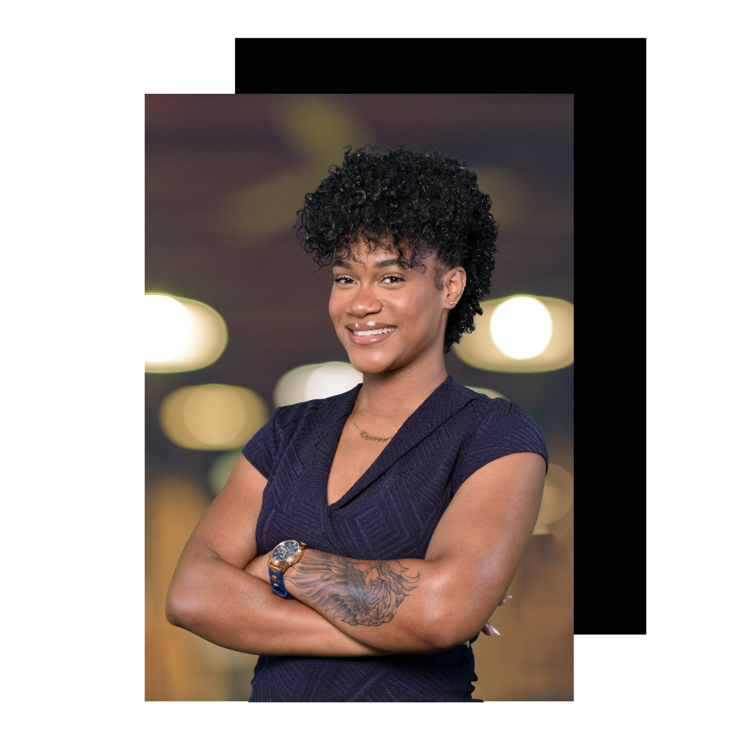 A smiling woman with short curly black hair wearing a dark blue dress, a watch, and a gold necklace, standing with her arms crossed against a blurred background of warm bokeh lights.