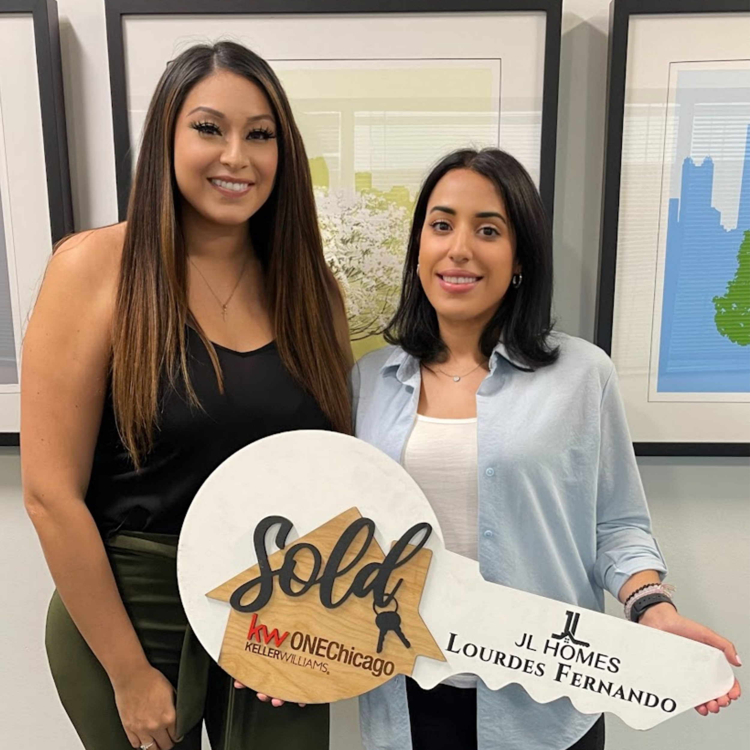 Two women standing together holding a large key-shaped sign that says 'SOLD' in black lettering. The sign features the logos of Keller Williams and HomeLourdes Fernanda, indicating a real estate transaction. They are smiling indoors with framed artwork in the background.