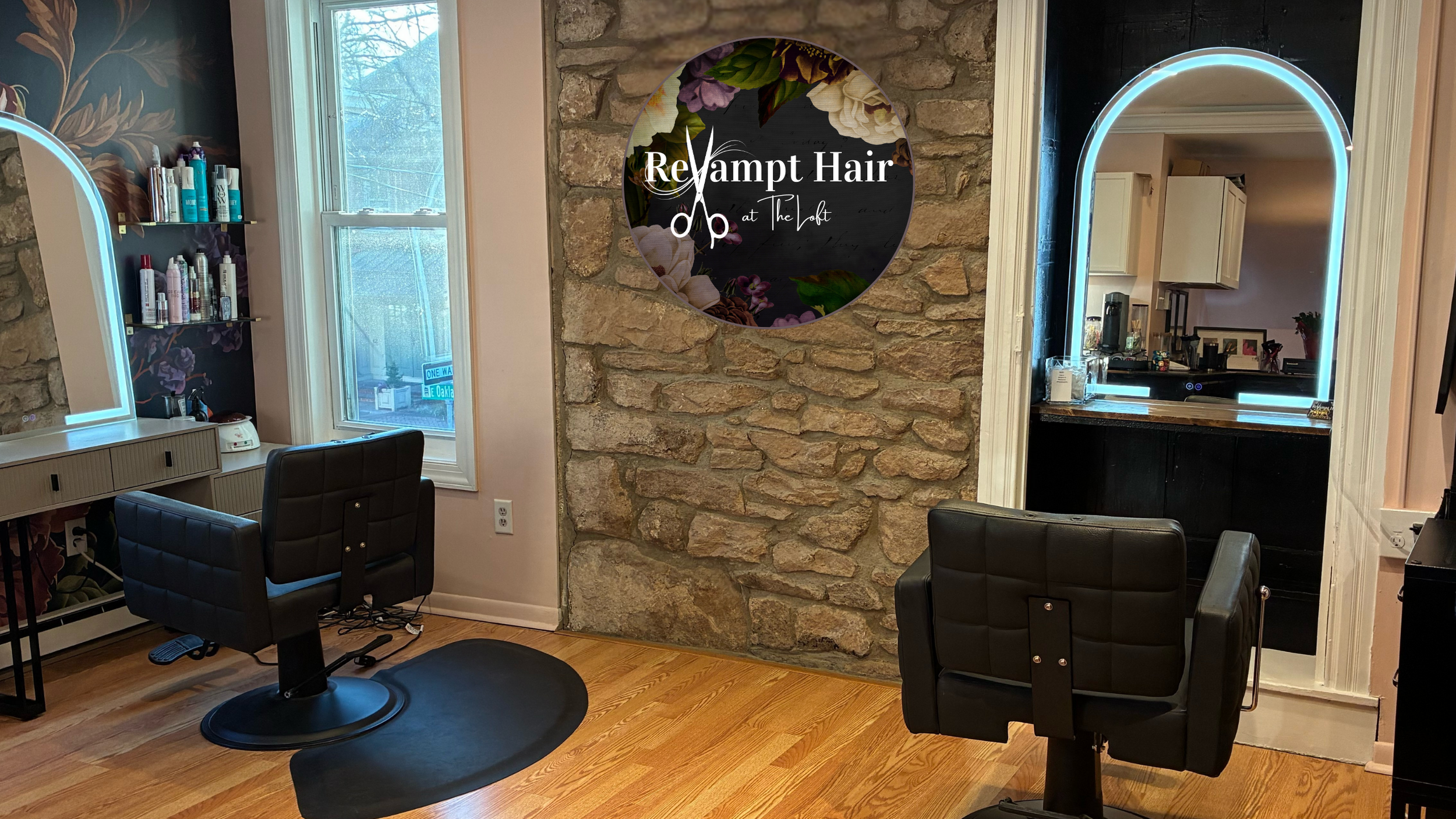 Interior of Revampt Hair salon in Doylestown, PA, with two black styling chairs, a window, a wall-mounted shelving with hair products, and a stone accent wall with a round sign reading 'Revamp Hair at The Loft'.