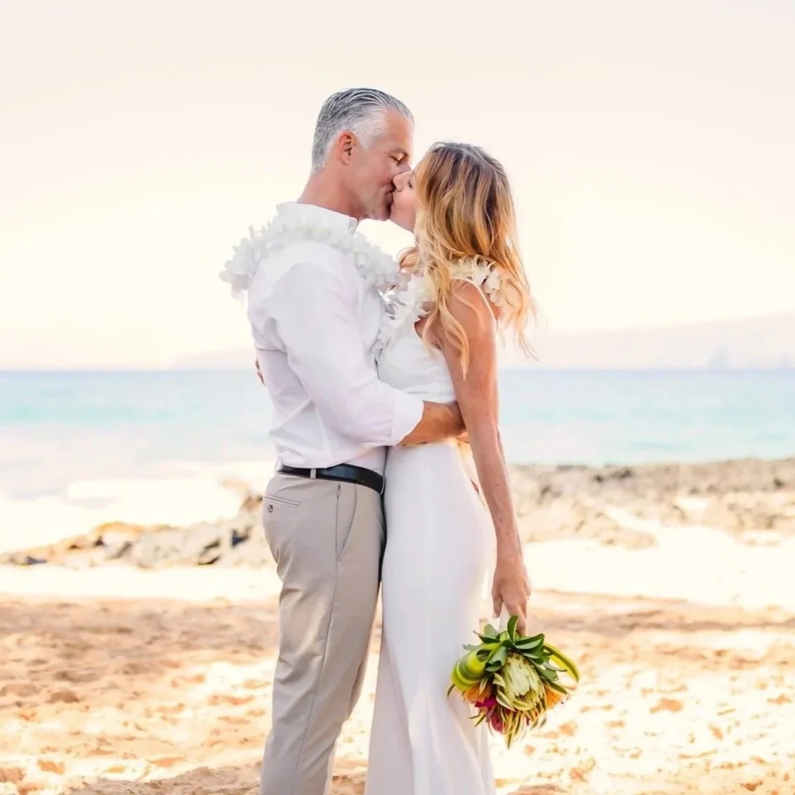 A couple on the beach kissing, with the woman holding a bouquet of flowers and the man in light-colored pants and a white shirt.