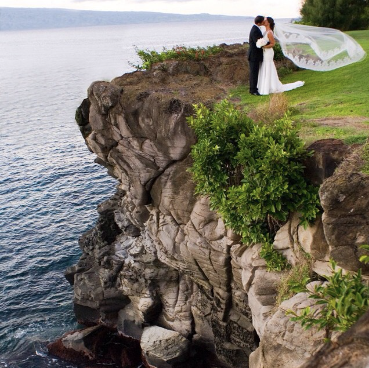 A newlywed couple in wedding attire, with the bride in a white gown and the groom in a black suit, embracing and smiling in a lush, green forest setting.