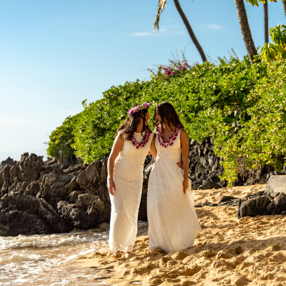 Two women dressed in white wedding dresses walking on a sandy beach with green bushes and palm trees behind them, under a blue sky, celebrating a wedding or special occasion.